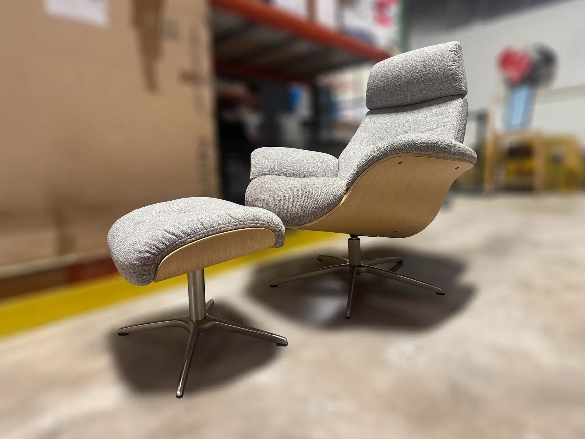 A modern light gray fabric lounge chair with a wood veneer shell and matching ottoman on an industrial floor.