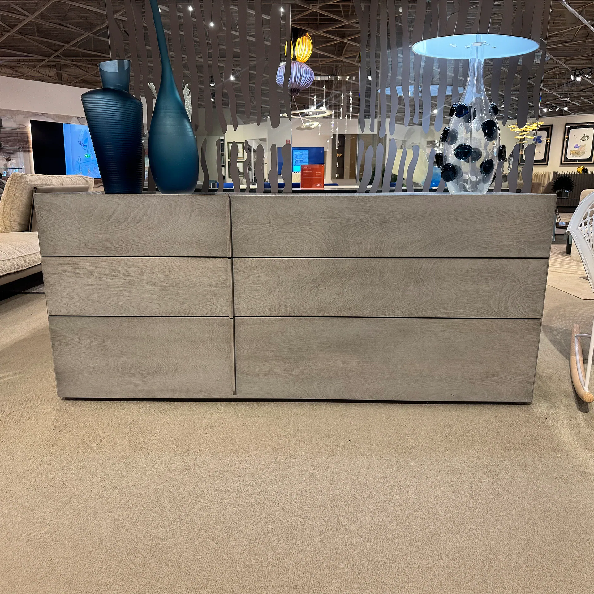 A light gray wooden dresser with six drawers, topped with three decorative vases: dark blue, teal, and clear glass with blue accents. The dresser sits on a beige carpet in a showroom setting.