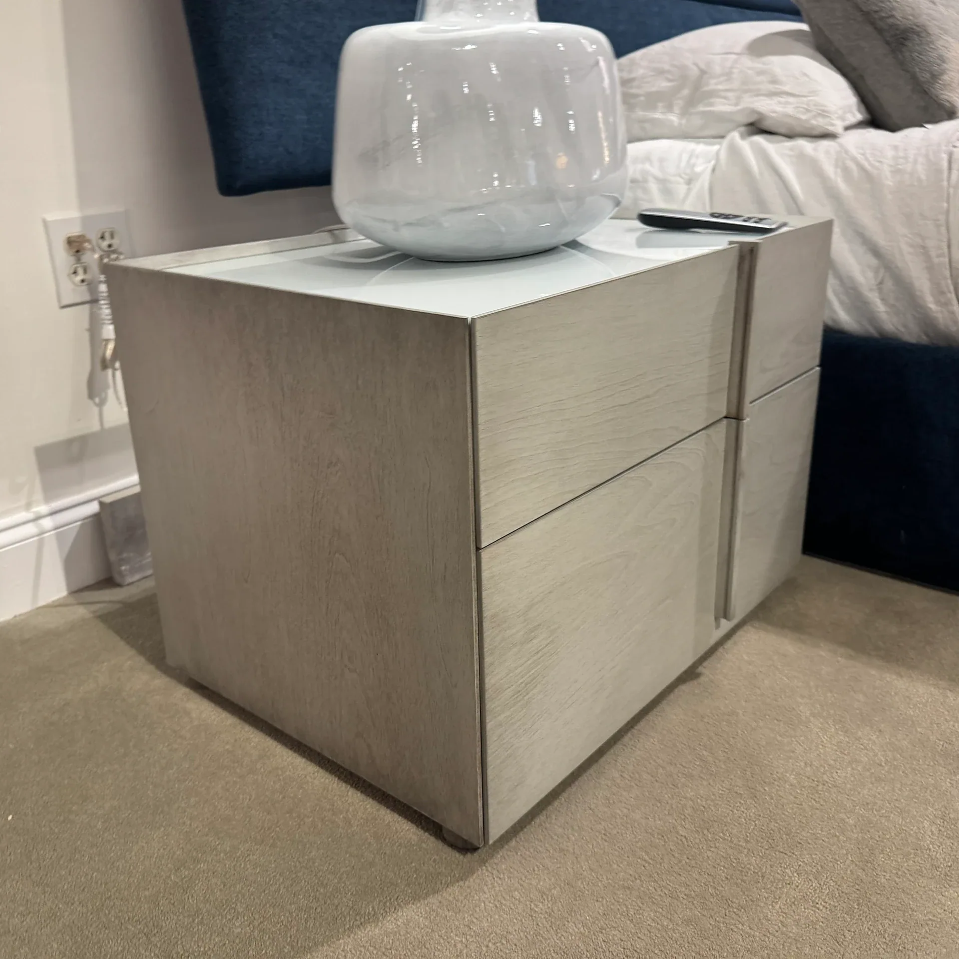 A light wood nightstand with a white glass top, a white marbled vase, and a remote control, next to a bed with a blue headboard and white bedding.