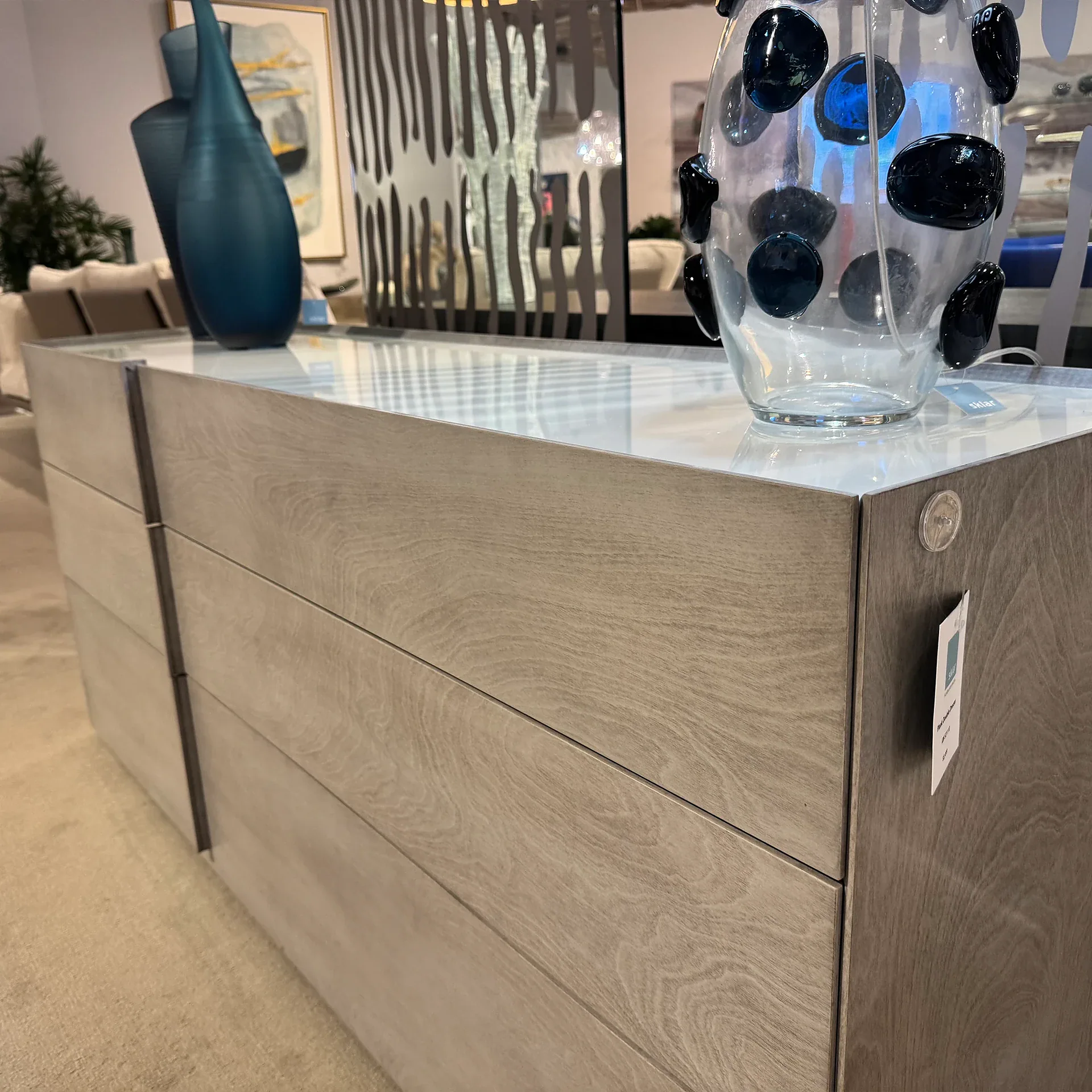 A modern light gray wooden dresser with a white top, featuring two tall teal vases and a clear glass vase with dark blue embellishments.