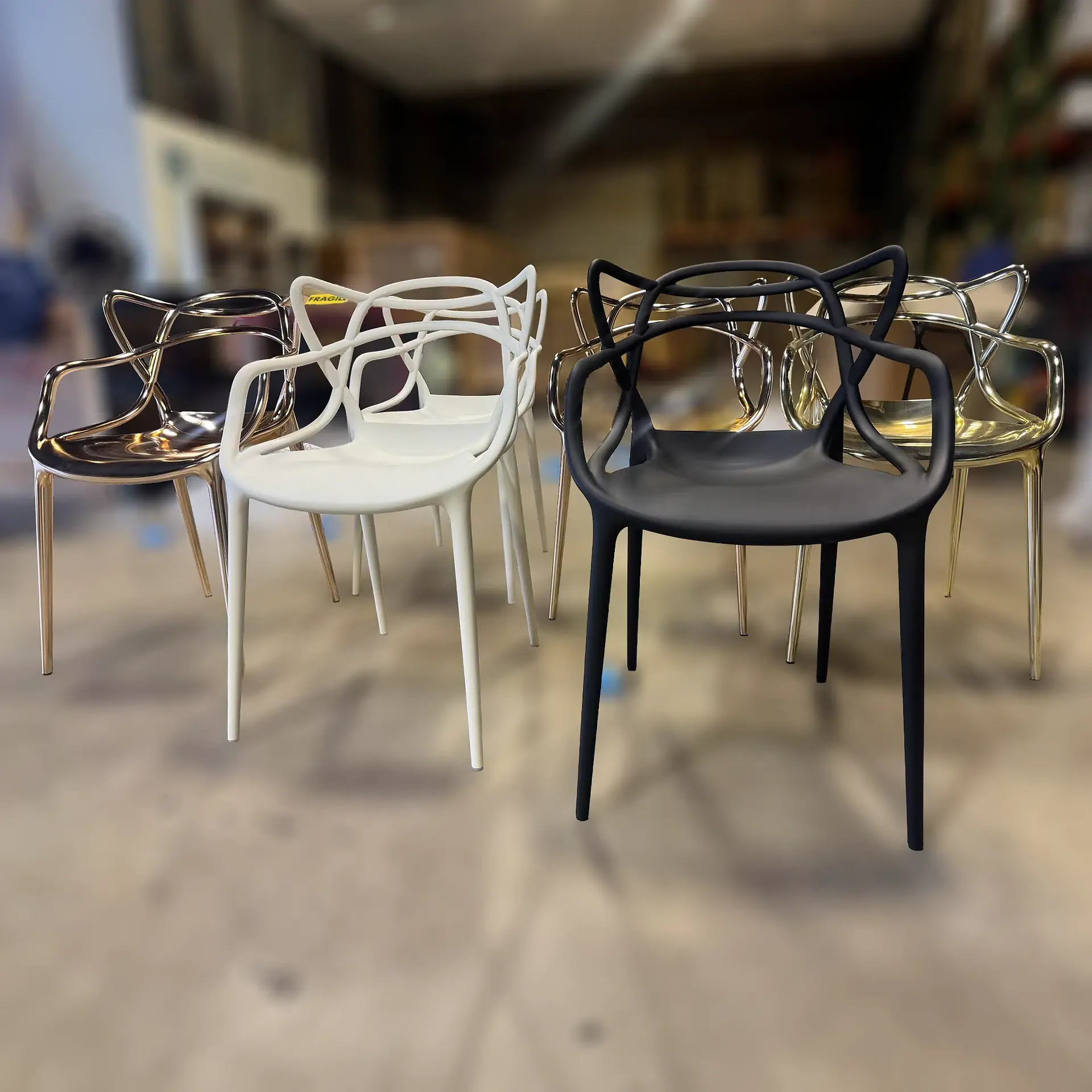 Six modern chairs with intricate, open-work backs and armrests. Three are shiny gold, one is matte white, and one is matte black. They are arranged on a light floor with a blurred background.