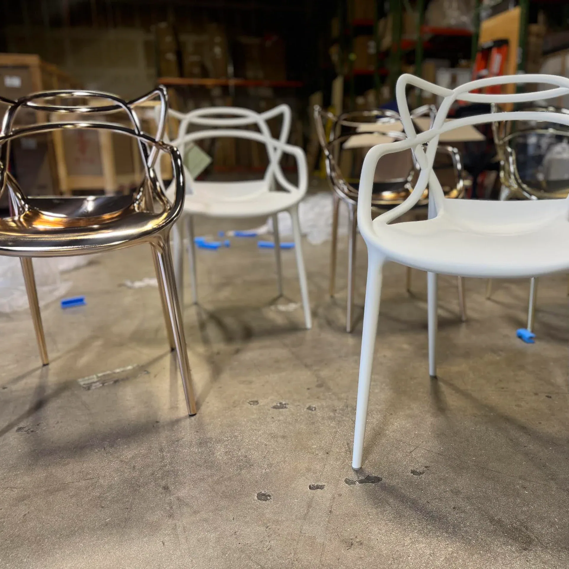Four modern chairs, two metallic gold and two matte white, with sculptural open backs, on a concrete floor in a warehouse.