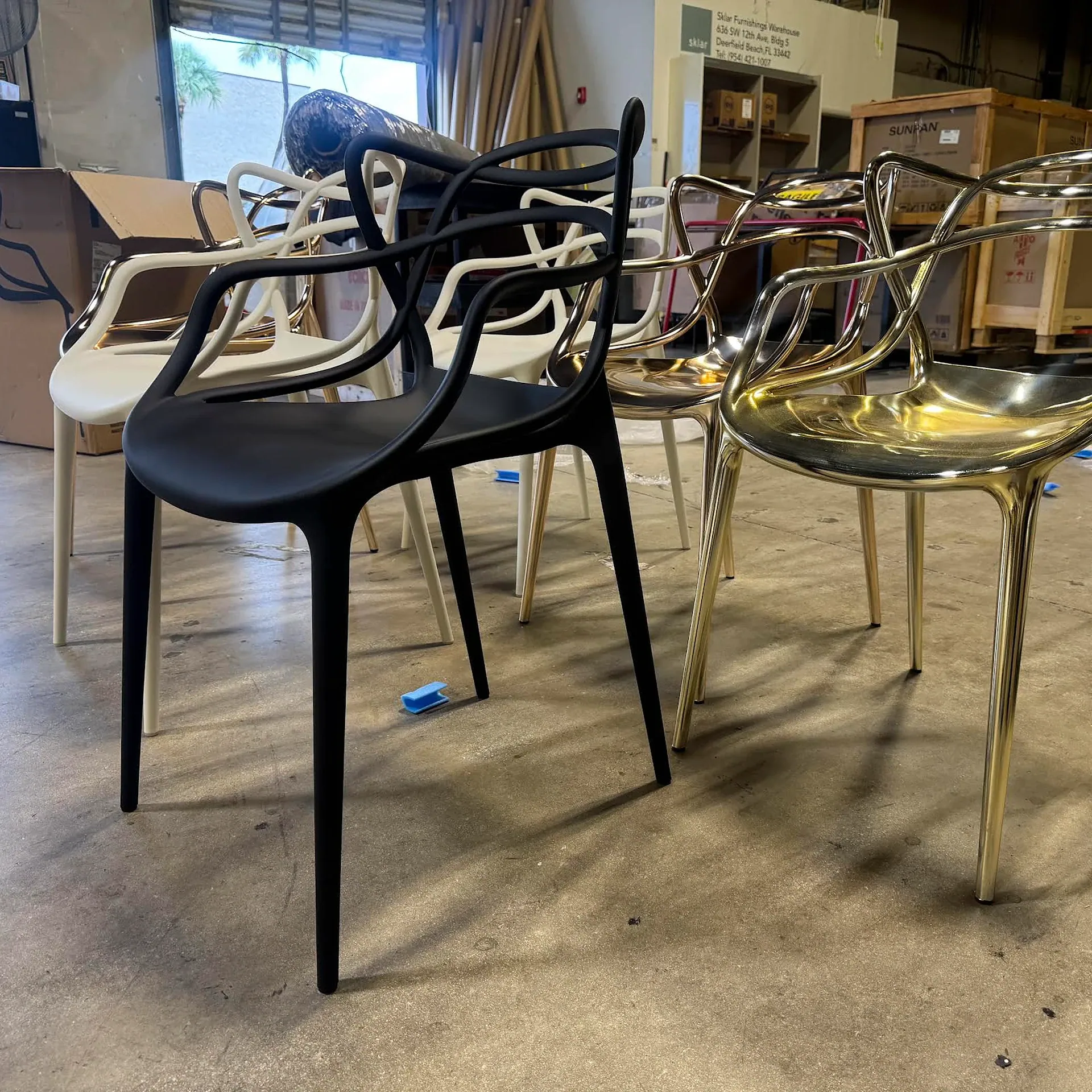 Modern sculptural chairs in matte black, off-white, and shiny gold finishes arranged on a concrete floor in a warehouse setting.