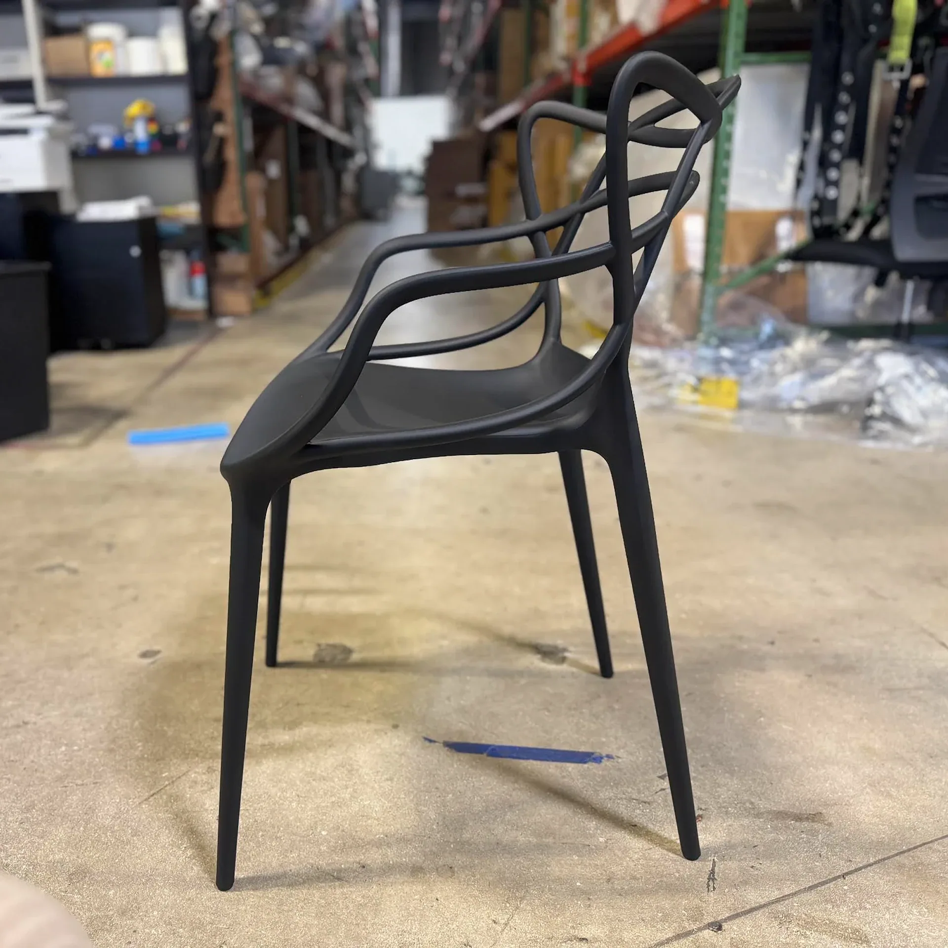 A matte black modern chair with a curved, abstract backrest and slender legs on a concrete floor in a blurry warehouse setting.
