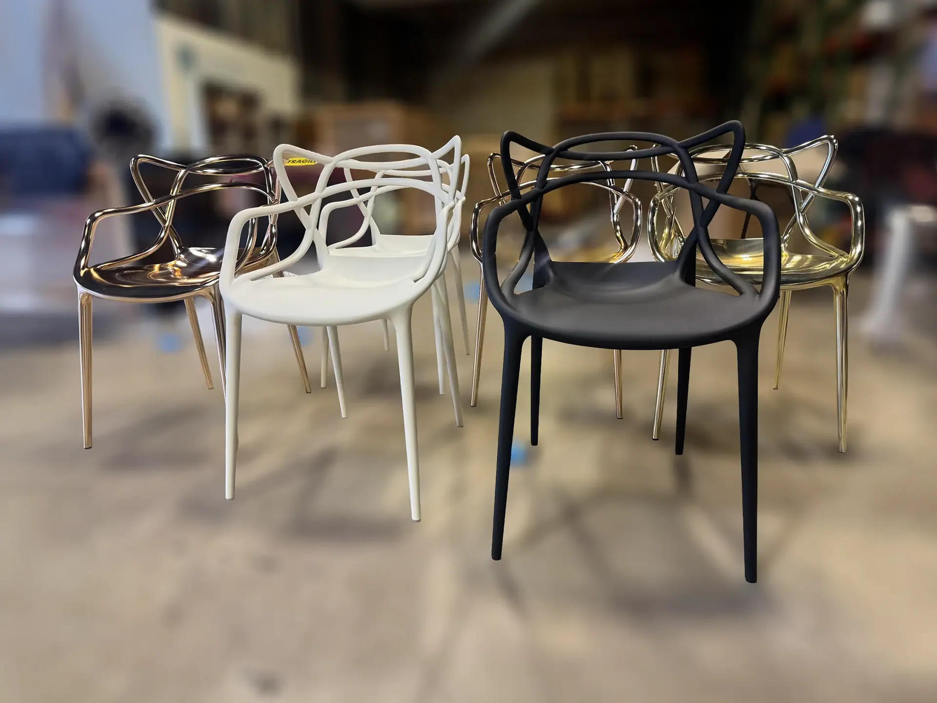 Eight modern chairs with sculptural backrests, arranged in two rows. Two white, two black, and four translucent gold chairs are visible on a light floor with a blurred background.