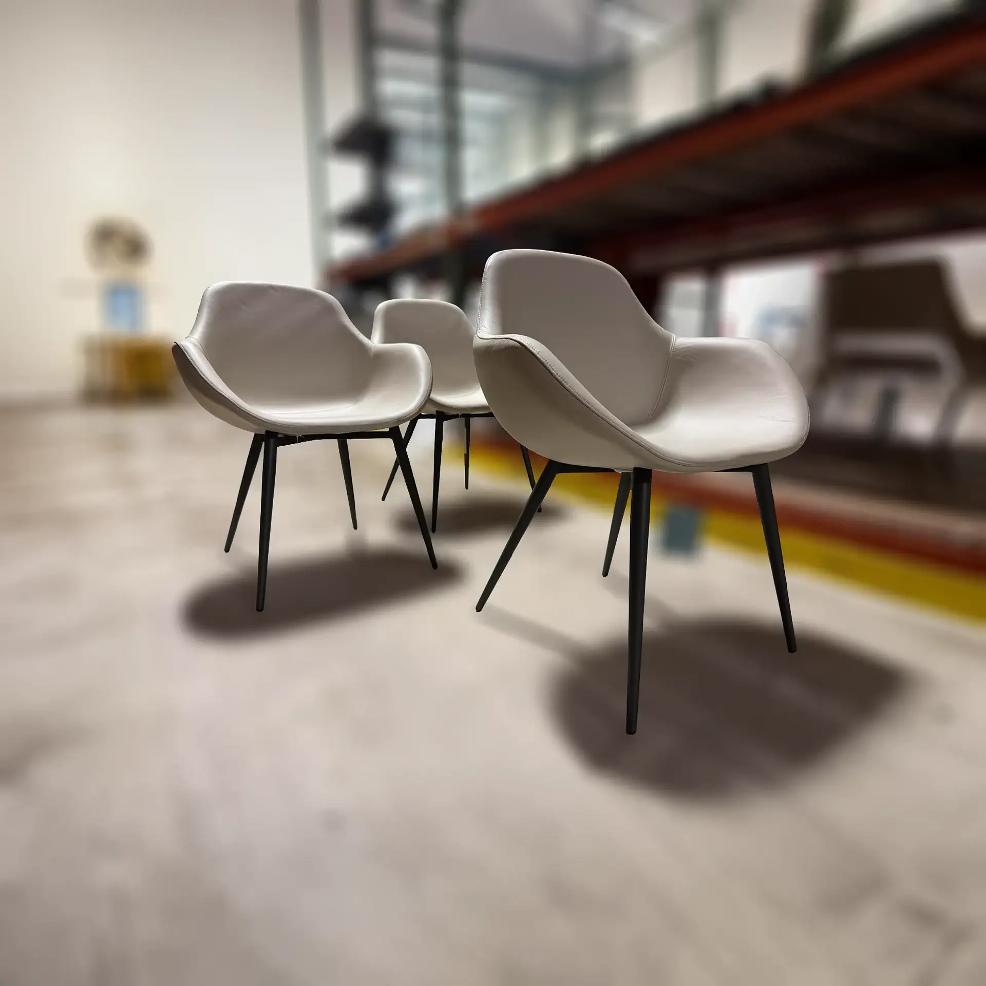 Three light beige modern chairs with black legs on a light floor, with a blurred industrial background.