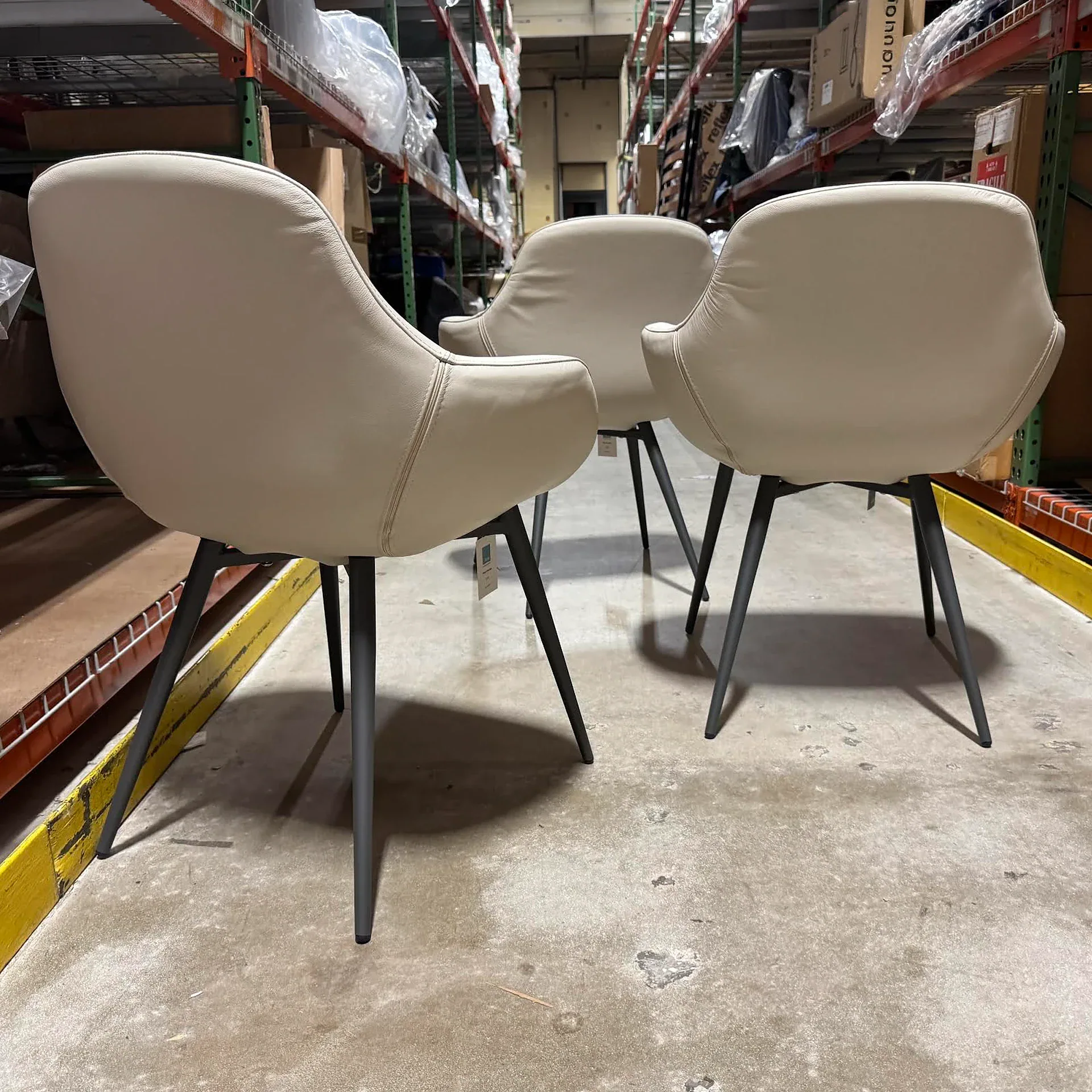 Three light beige upholstered chairs with dark metal legs, viewed from behind in a warehouse setting with industrial shelving in the background.
