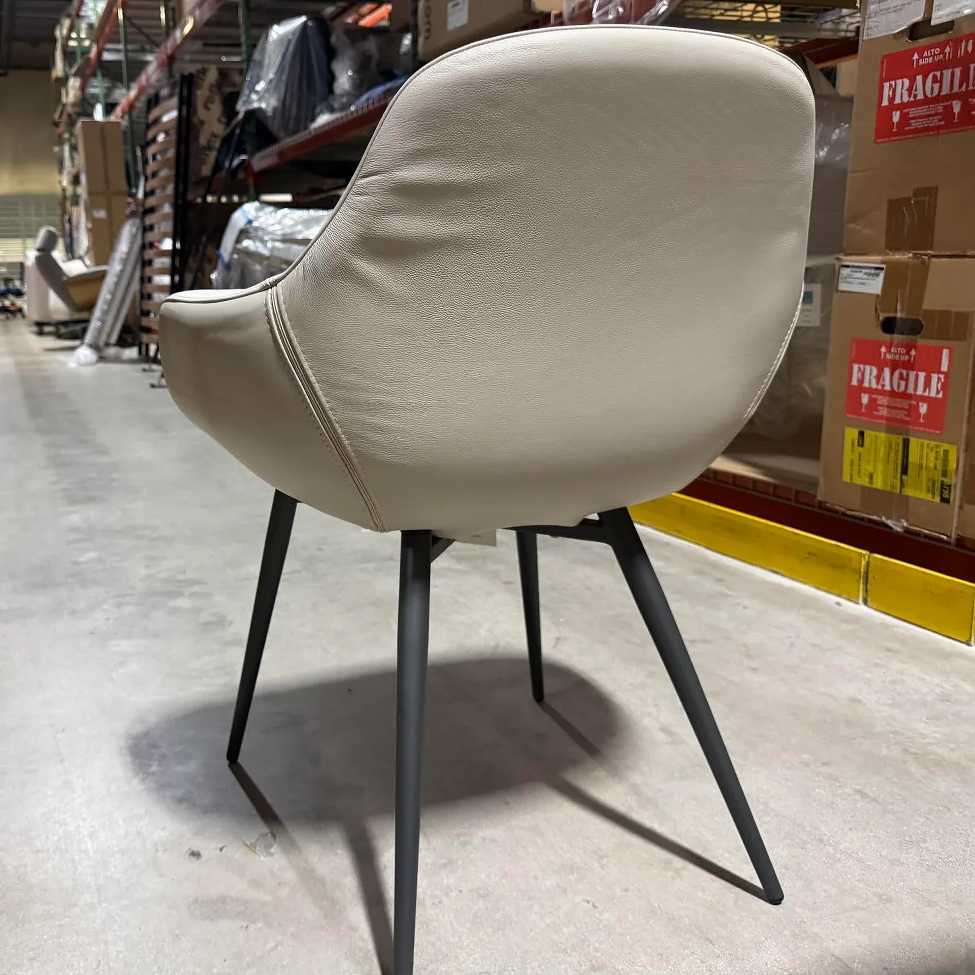 A light beige upholstered chair with black tapered legs stands on a concrete floor in a warehouse.