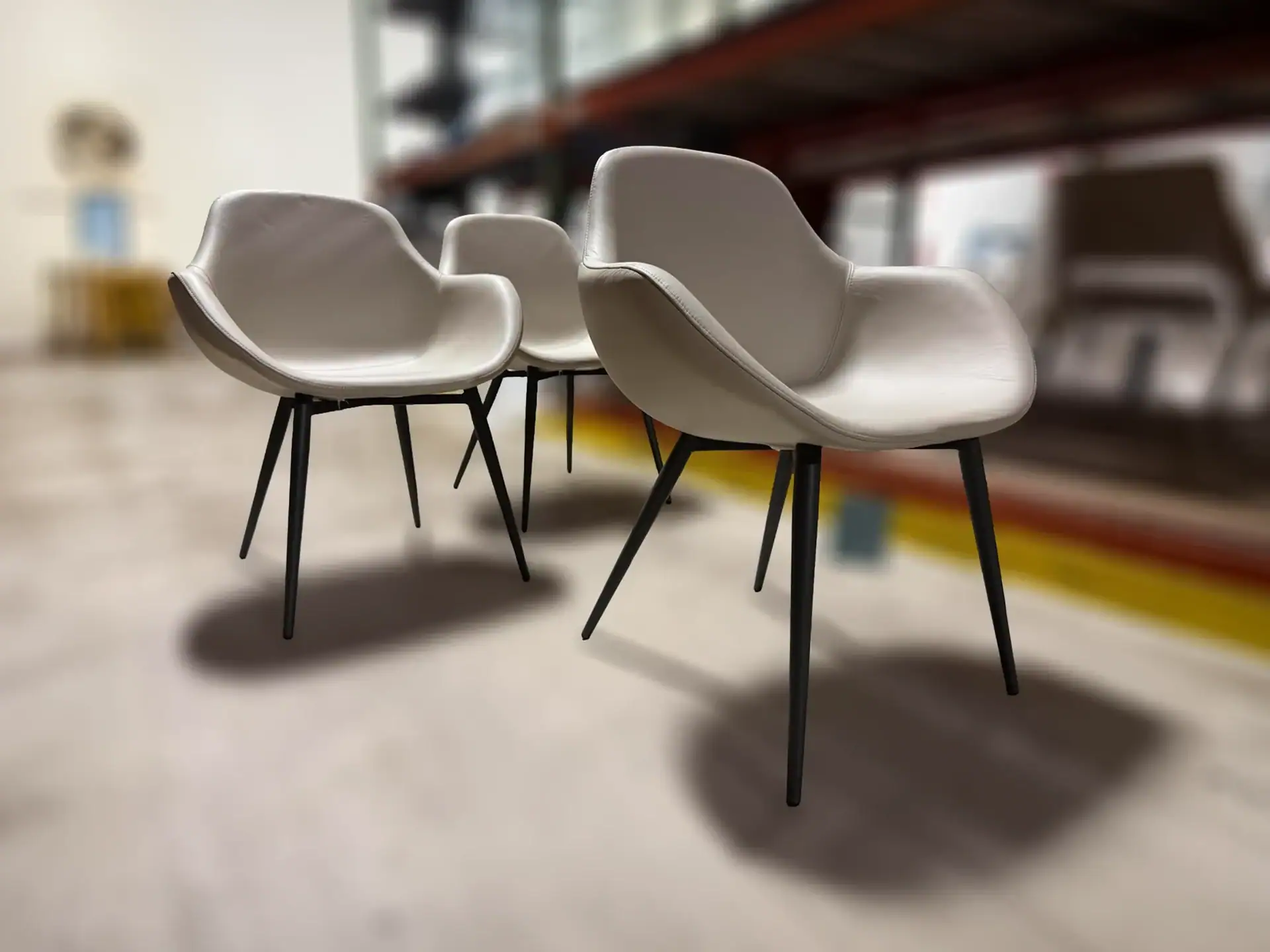 Three light beige modern chairs with black legs on a light floor, with a blurred warehouse background.