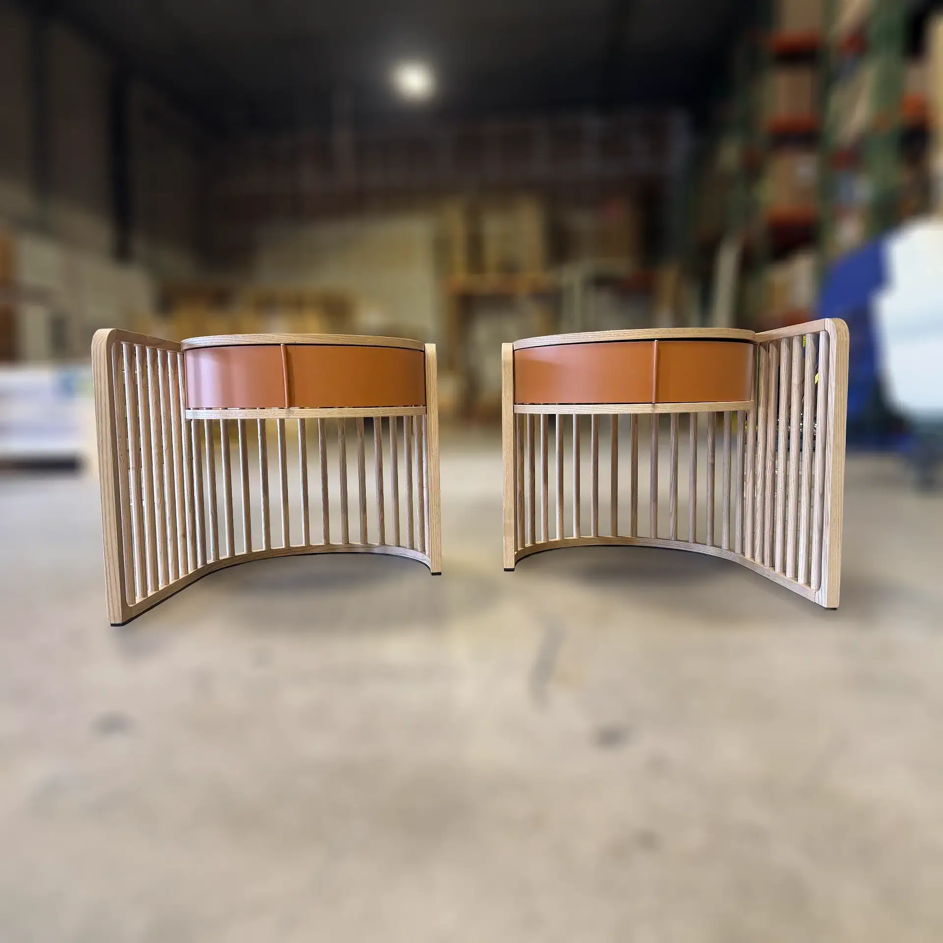 Two identical curved wooden side tables with vertical slats and reddish-brown leather tops, set against a blurred industrial background.