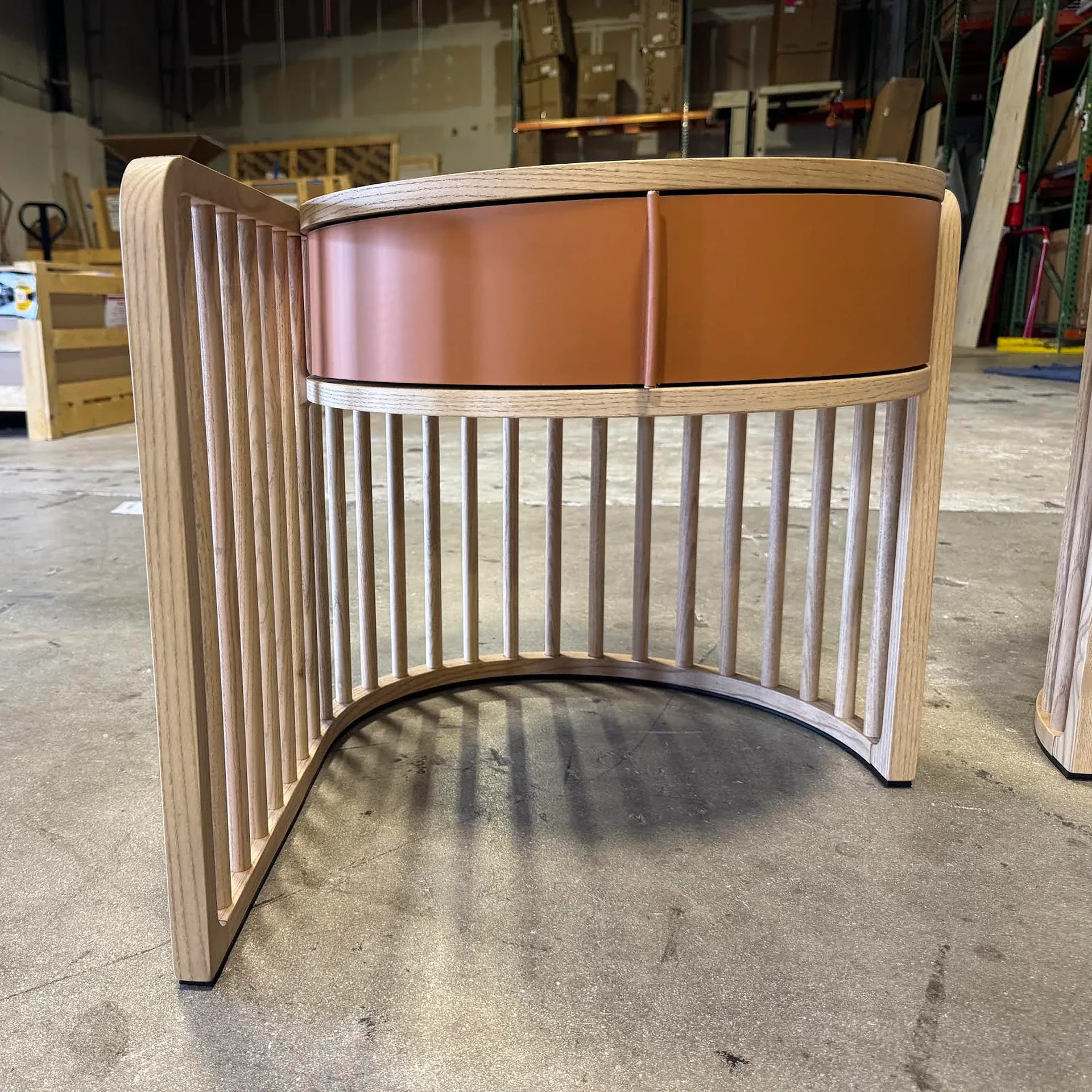 A curved wooden side table with vertical slats and a reddish-brown drawer, standing on a concrete floor in a warehouse setting.