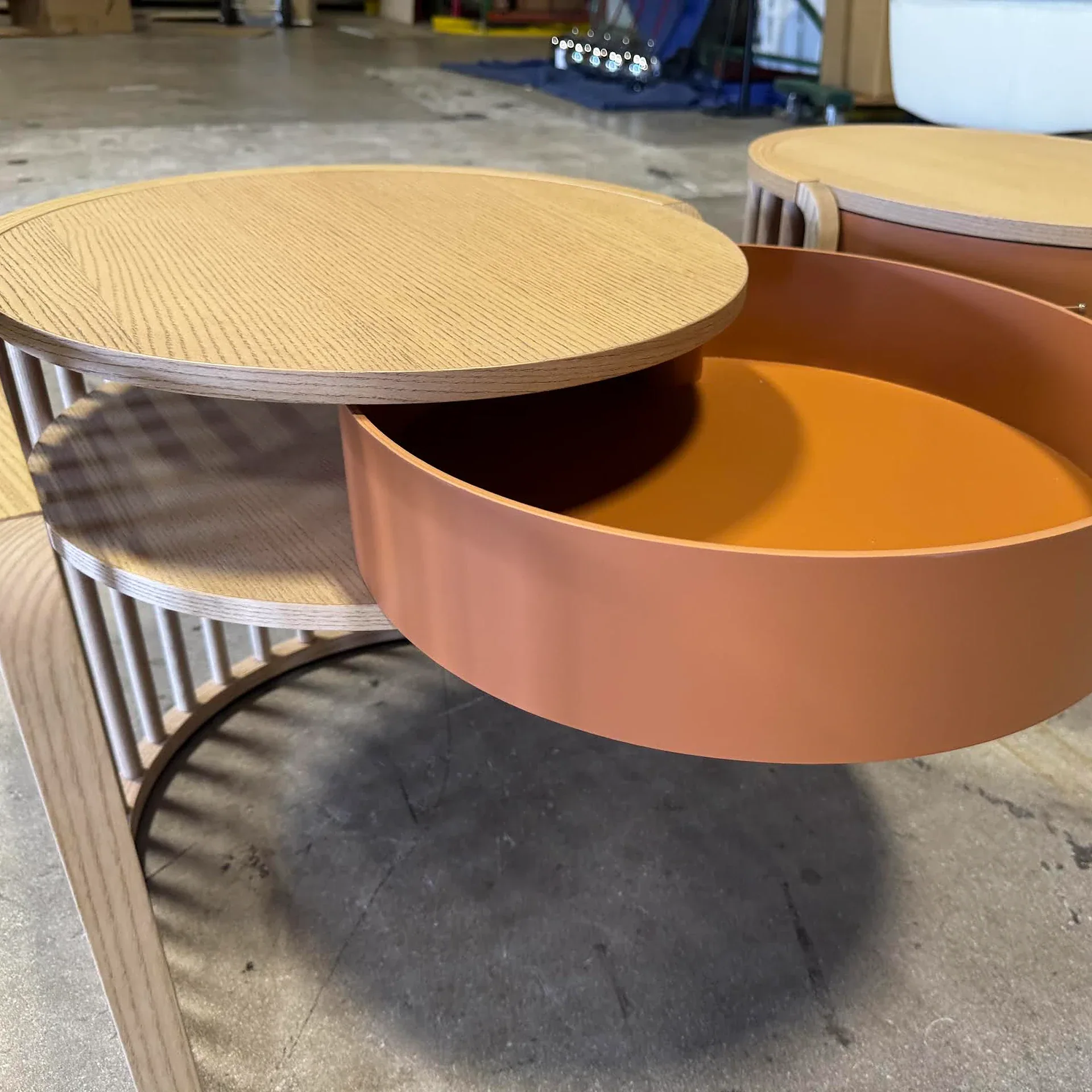 A modern, multi-tiered round wooden table with a terracotta-colored pull-out drawer, set on a concrete floor.