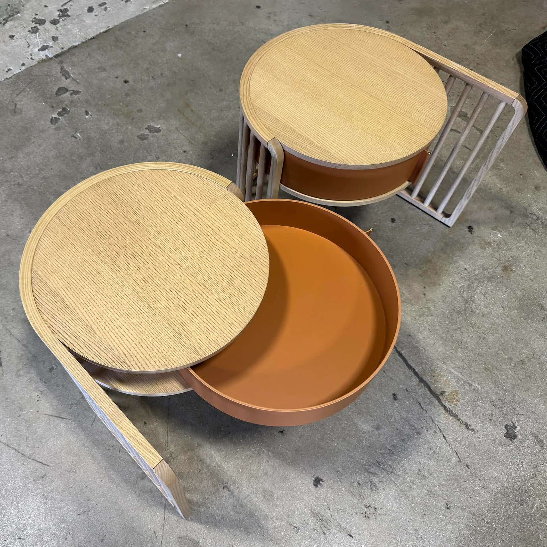Two modern round side tables with light wood tops and terracotta storage compartments on a concrete floor. One table is open, revealing the storage.