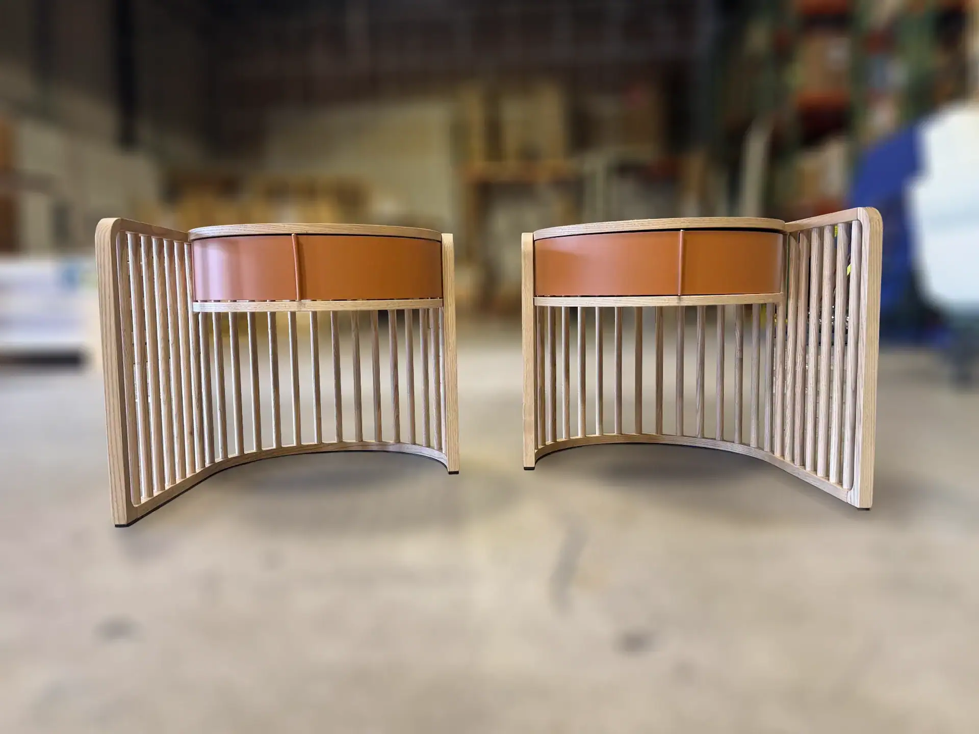 Two identical curved wooden side tables with slatted sides and brown leather-fronted drawers, set against a blurred workshop background.