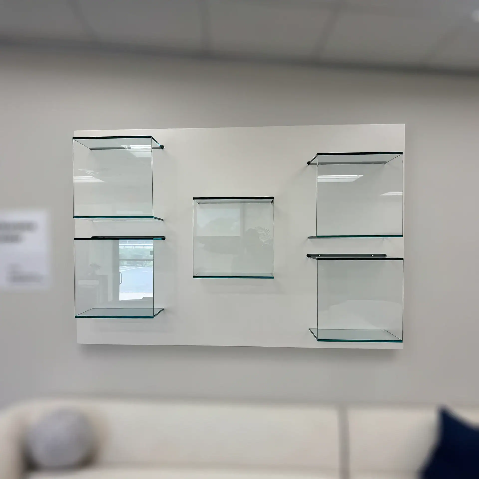 Five empty clear glass display shelves mounted on a white rectangular panel on a light gray wall, with a blurred sofa in the foreground.