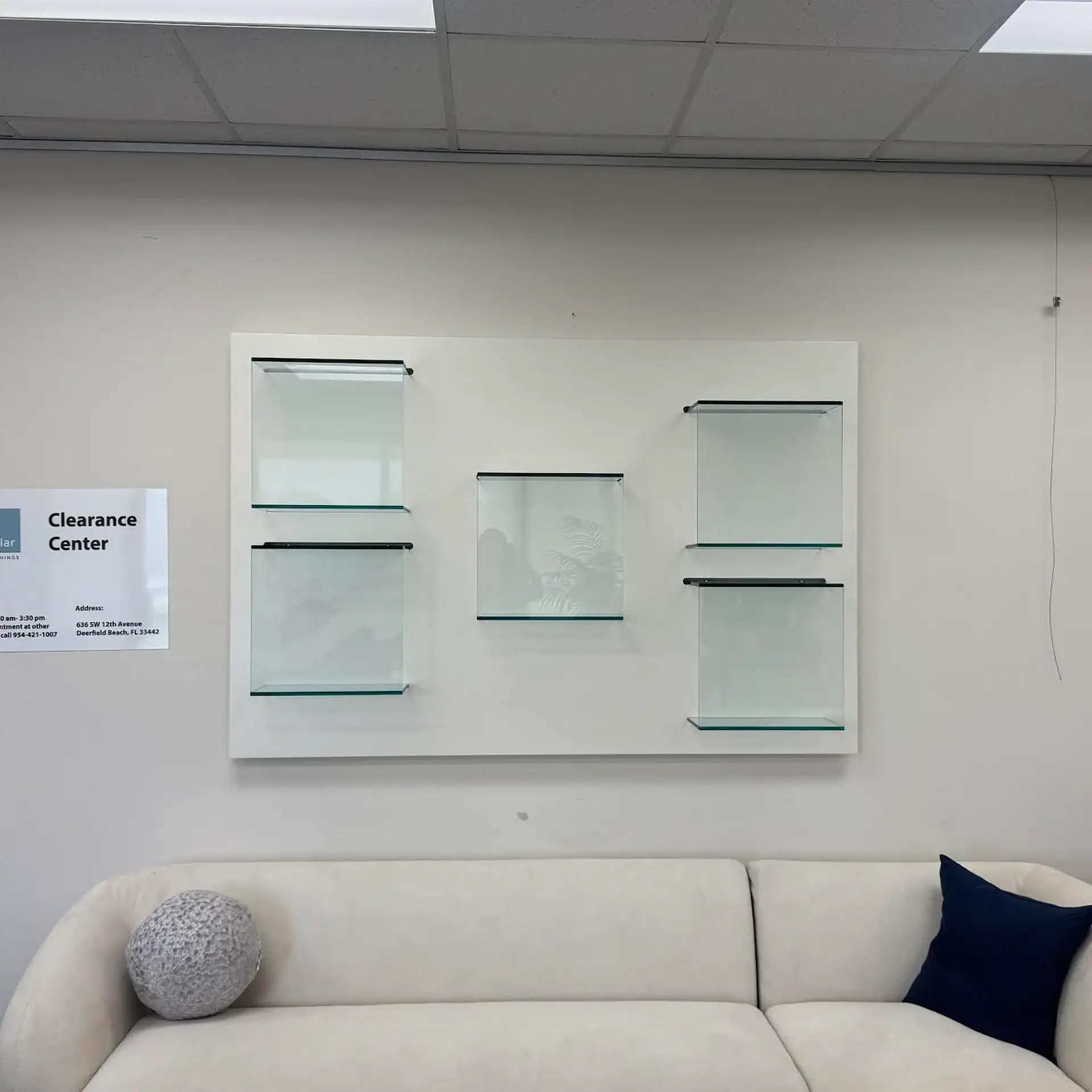 A white sofa with a grey textured cushion and a blue cushion sits below a wall-mounted display of five empty glass shelves. A 'Clearance Center' sign is visible on the left.