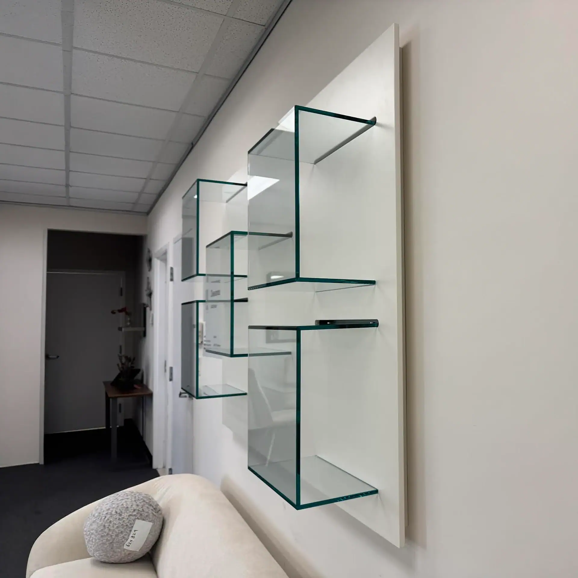 Modern glass shelves mounted on a white wall panel, with a cream sofa and grey spherical pillow in the foreground. An office hallway is visible in the background.