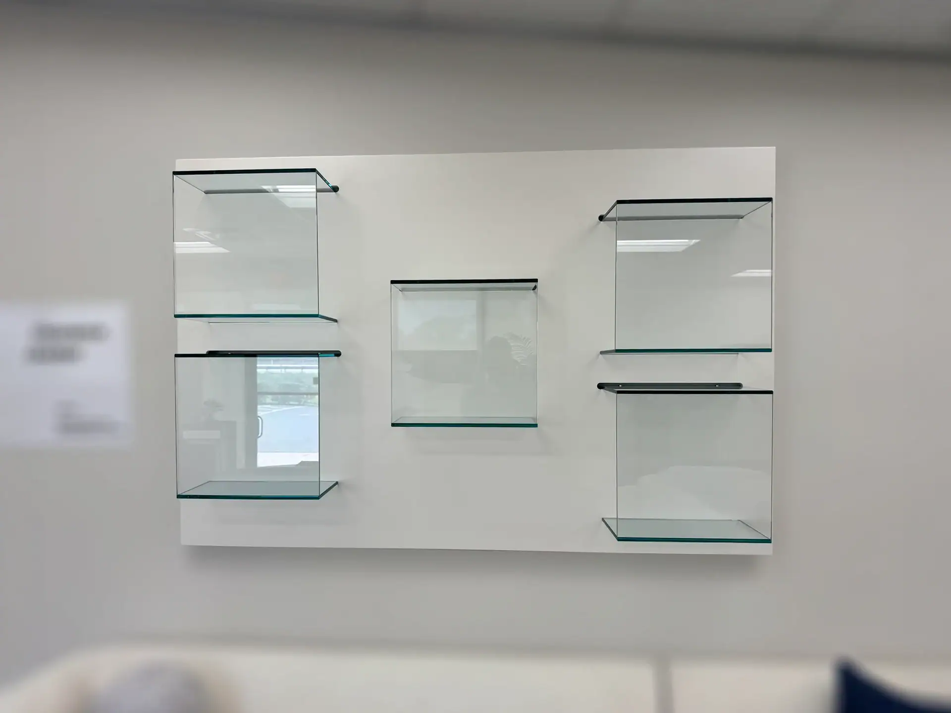 Five empty, clear glass display shelves with black brackets mounted on a white rectangular panel on a light gray wall. A blurred sofa is at the bottom.