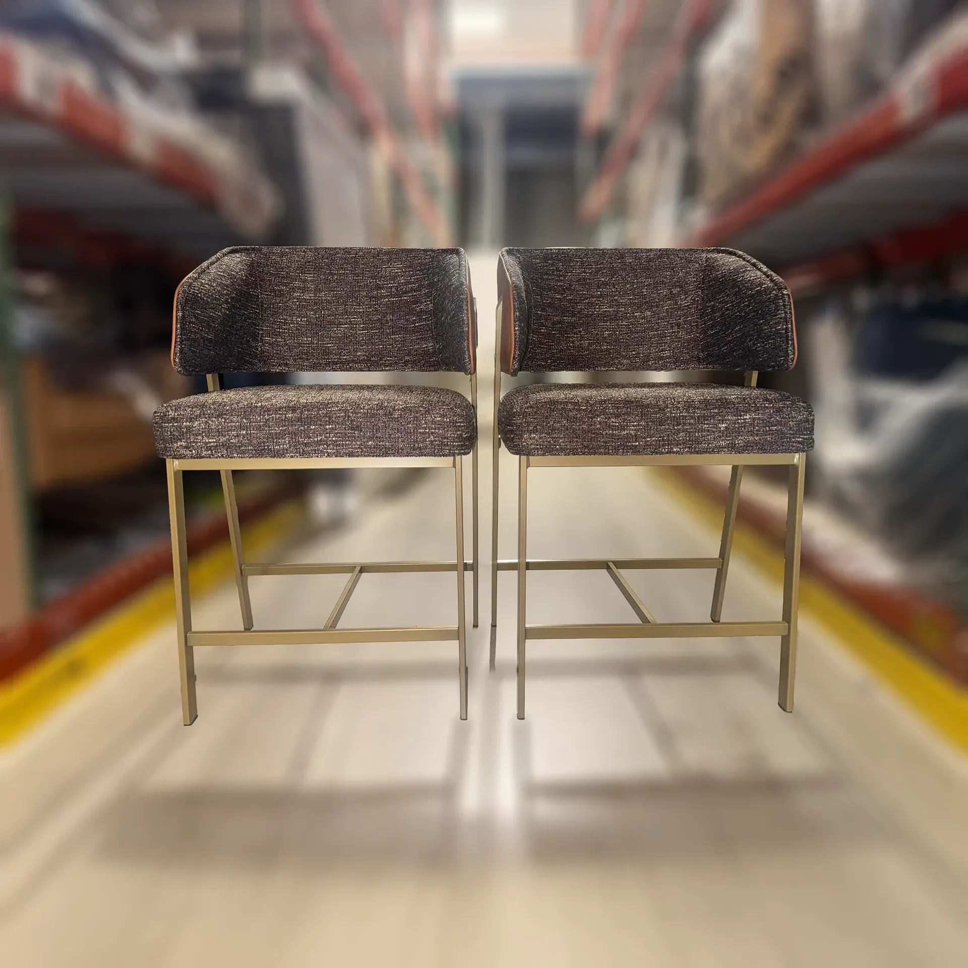 Two dark upholstered counter stools with gold metal frames stand side-by-side in a blurred warehouse setting.