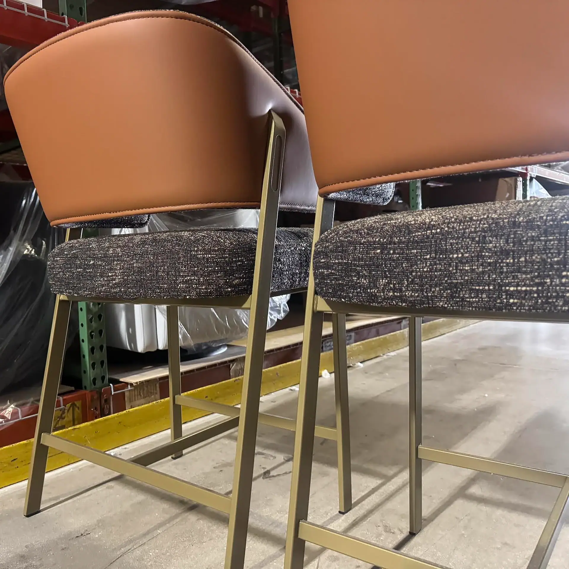 Two modern bar stools with tan backs, speckled dark fabric seats, and gold metal frames in a warehouse setting.