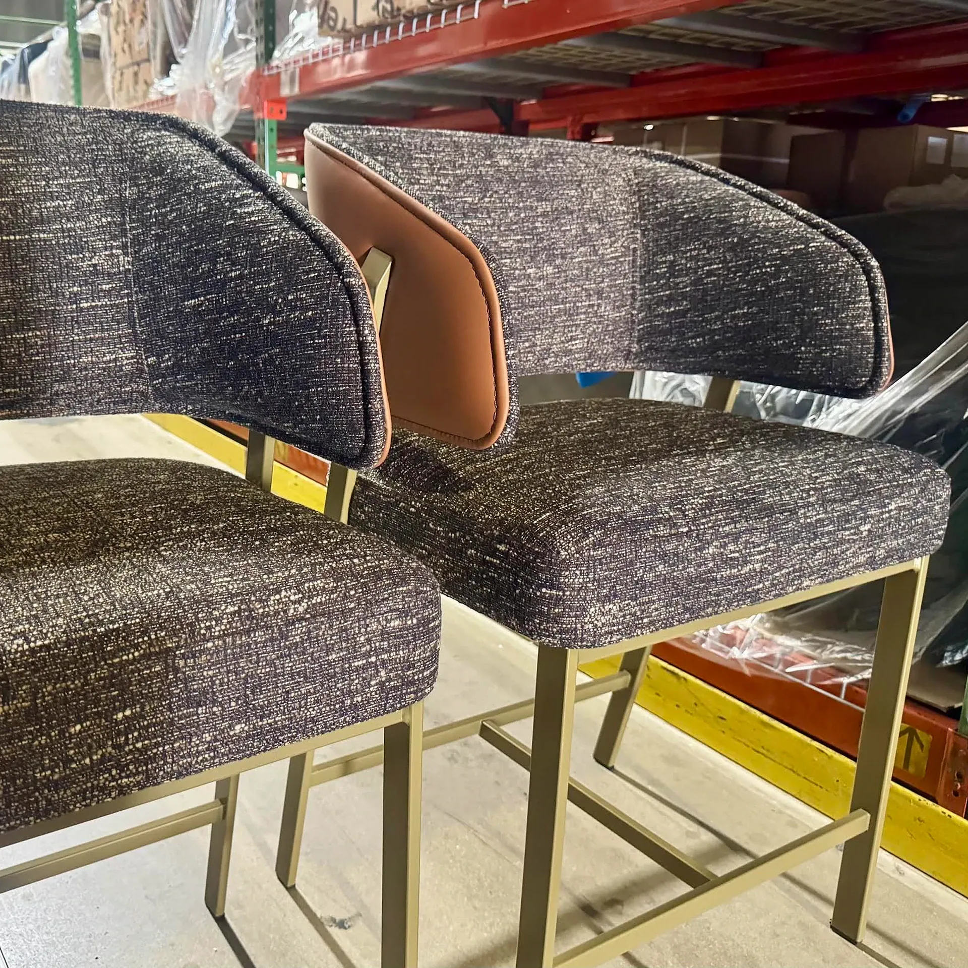 Two modern dark upholstered chairs with tan leather accents and gold metal frames in a warehouse setting.