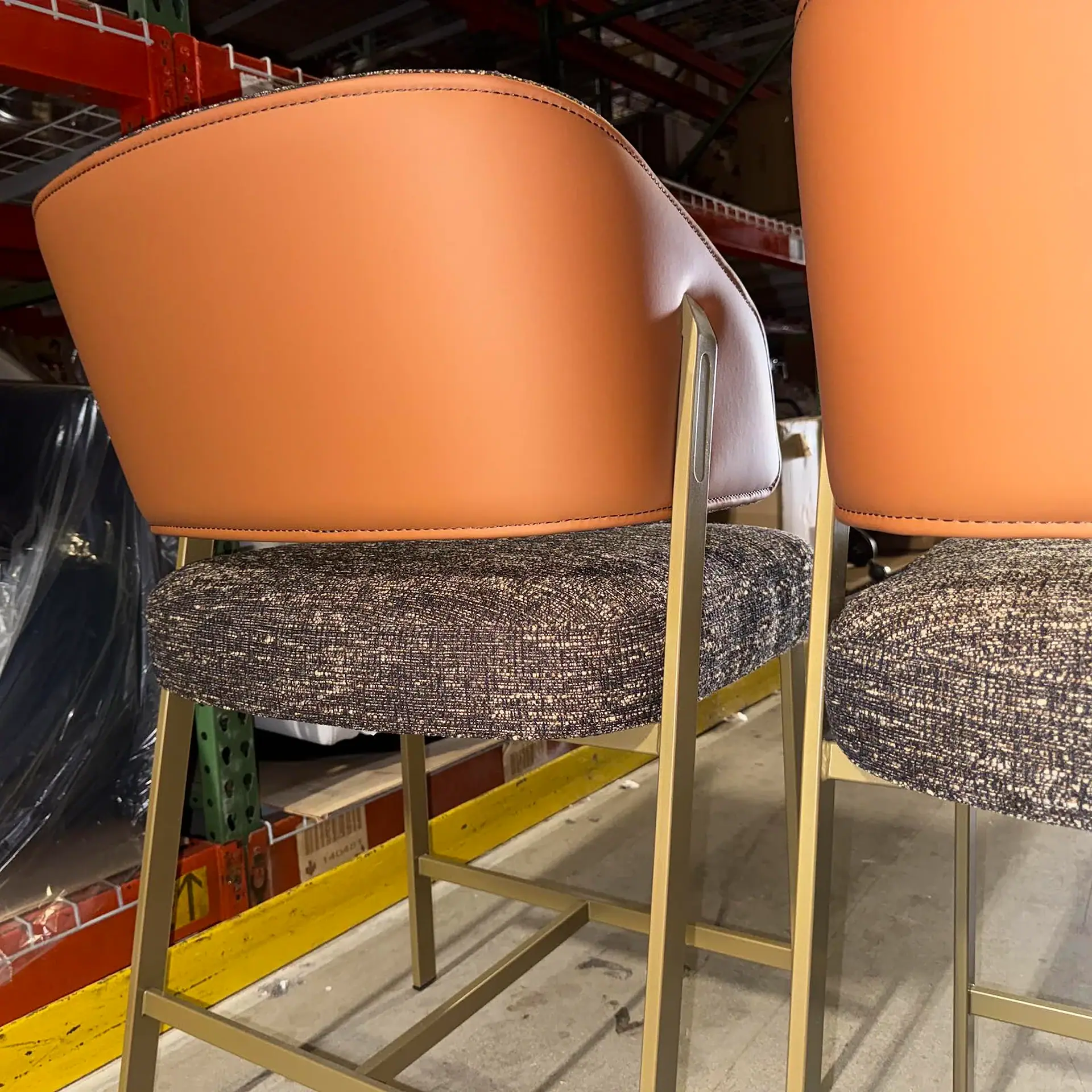 Two modern bar stools with cognac backrests, textured gray seats, and gold metal frames in a warehouse setting.