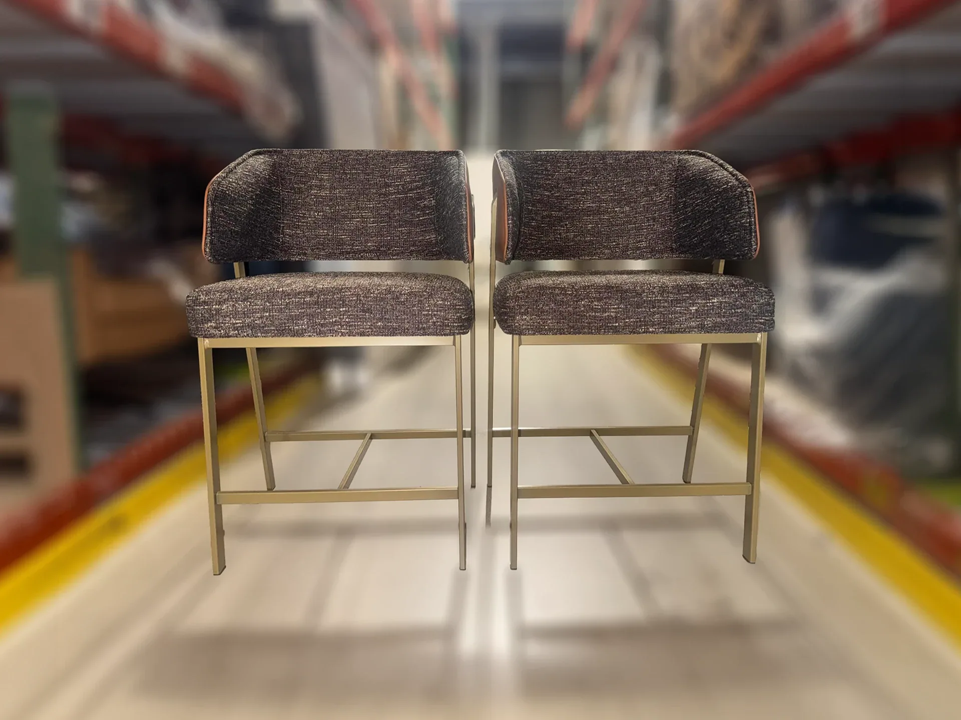 Two identical dark gray upholstered bar stools with gold metal frames stand side-by-side in a blurred warehouse setting.