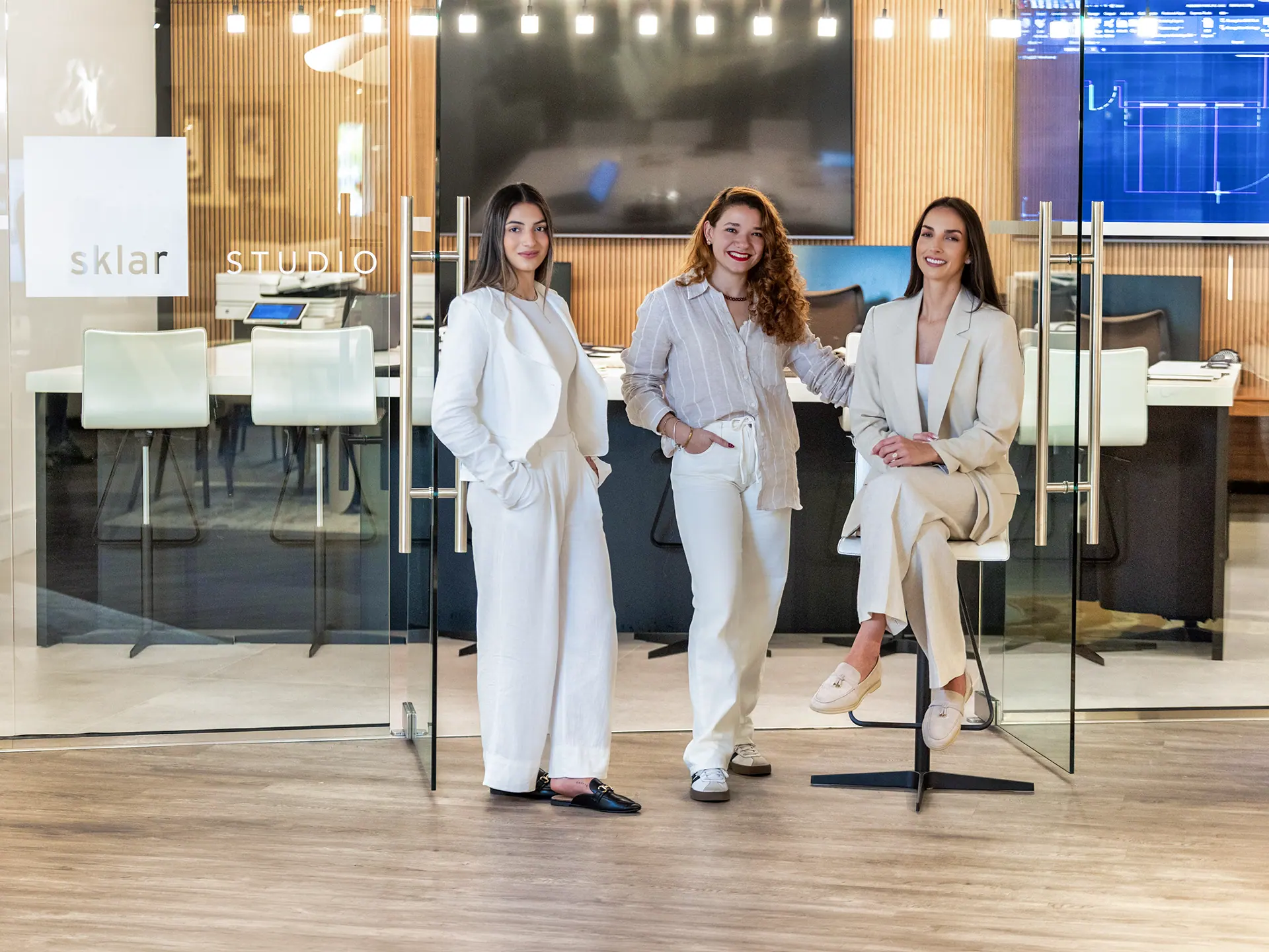 Three women in professional attire stand and sit in front of a modern office studio with 'sklar STUDIO' on the glass partition.