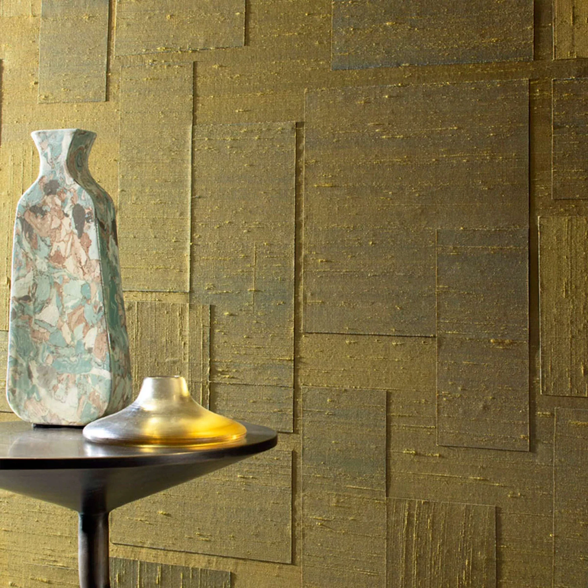 Vase and metallic dish on a dark table in front of a geometric gold wallpaper.