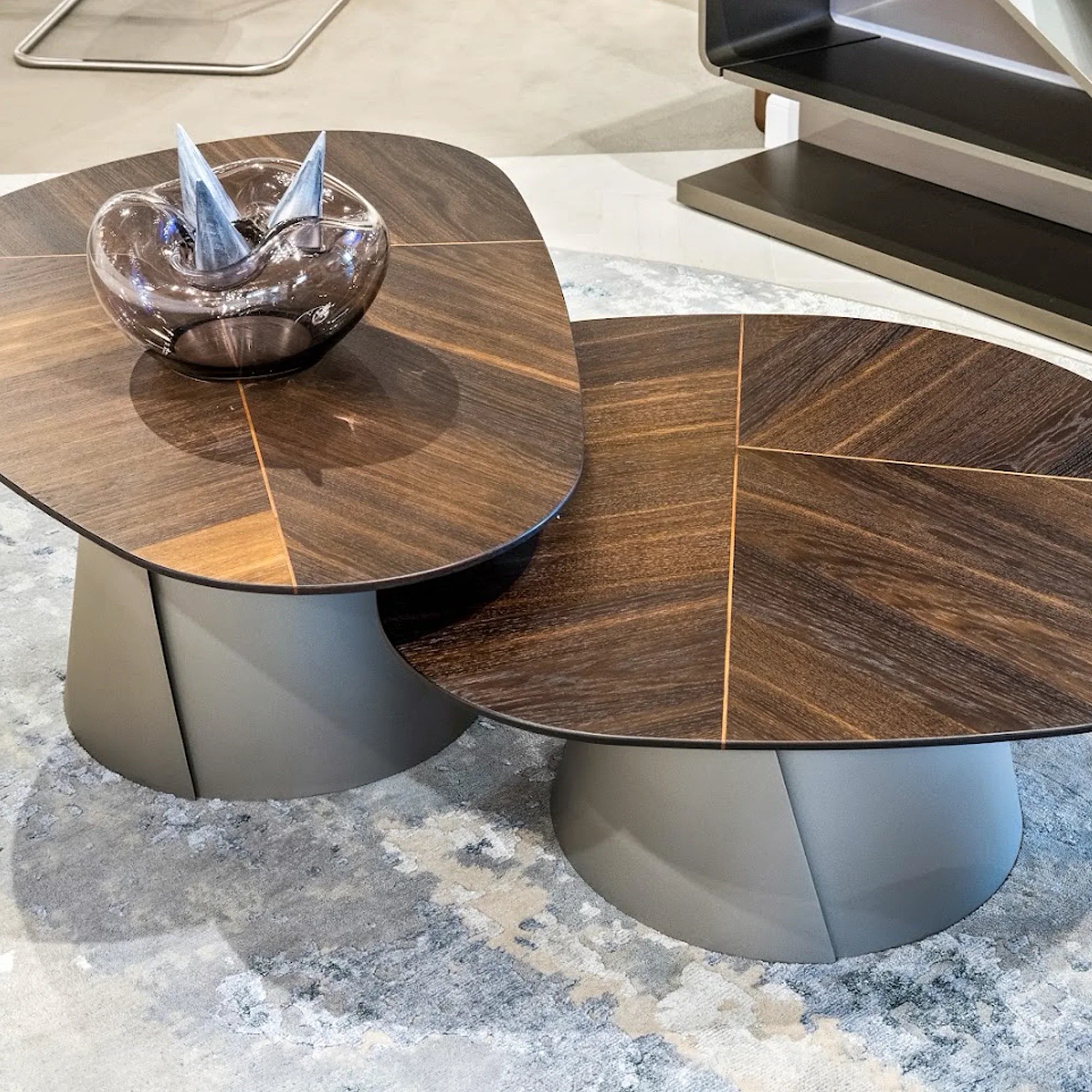 interior-designer-program-discounts Two modern, irregularly shaped coffee tables with dark wood tops and gray bases on a patterned rug, one holding a decorative glass bowl.