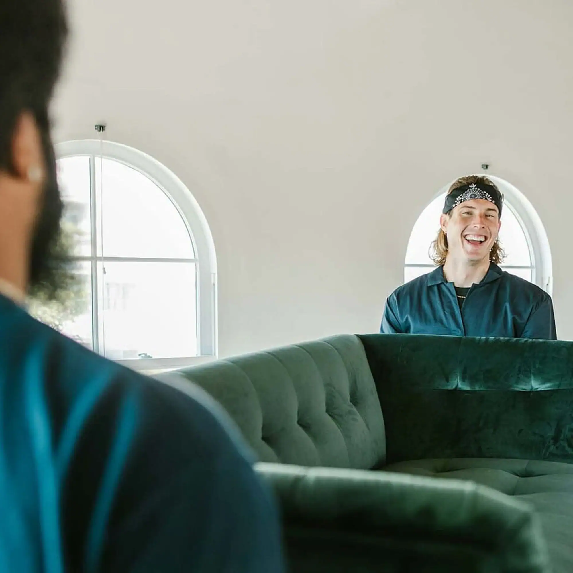 interior-designer-program-installation Two men moving a dark green velvet sofa in a bright room with arched windows. One man smiles from behind the sofa, while the other's back is to the viewer.