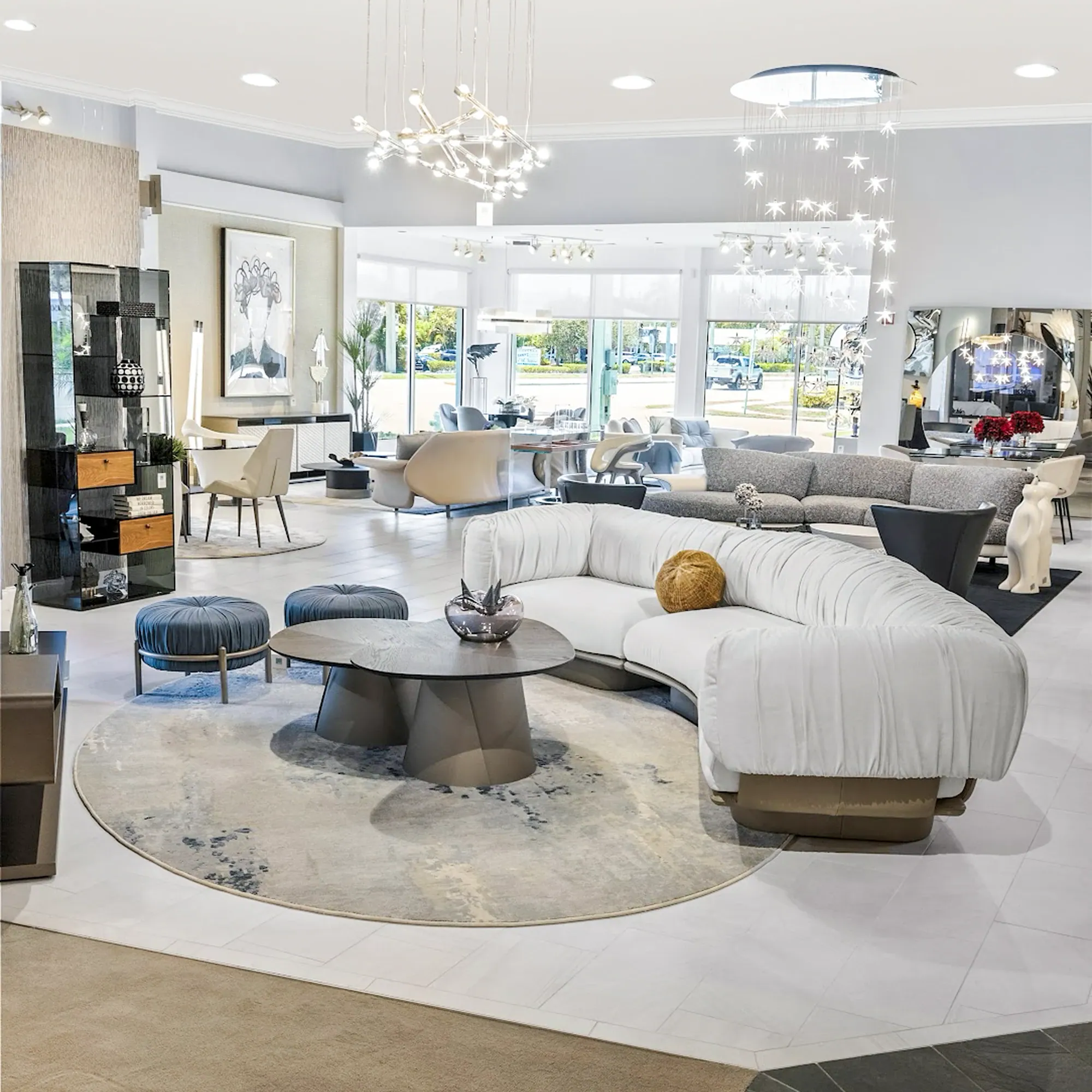 interior-designer-program-showroom A modern furniture showroom with a large, curved white sofa, a round coffee table, and two blue ottomans on a circular rug. Unique chandeliers hang from the ceiling, and large windows overlook a street scene.