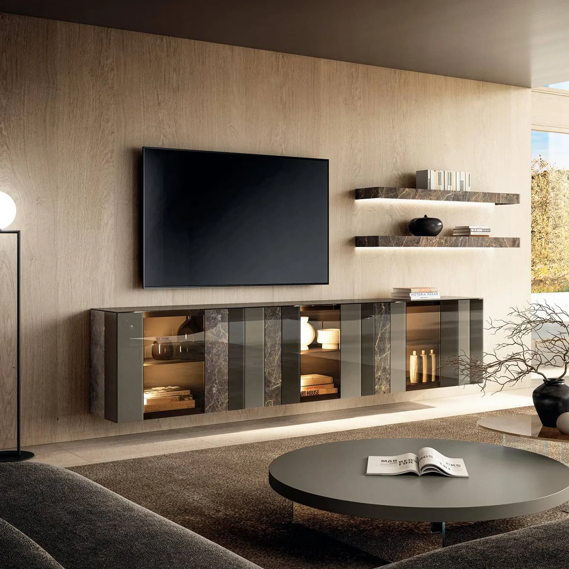 A modern living room with a wall-mounted TV, a floating dark gray media console with glass doors, and two illuminated marble-patterned shelves on a wood-paneled wall. A dark gray sofa and two round coffee tables with decor are in the foreground, with a window showing autumn trees on the right.