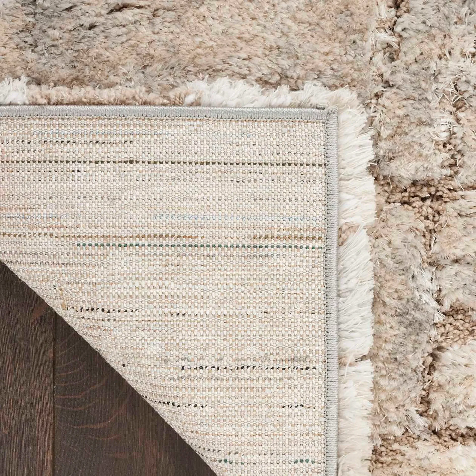 Overhead view of a light beige and cream shaggy rug, with one corner folded back to reveal its woven backing and a dark brown wooden floor.