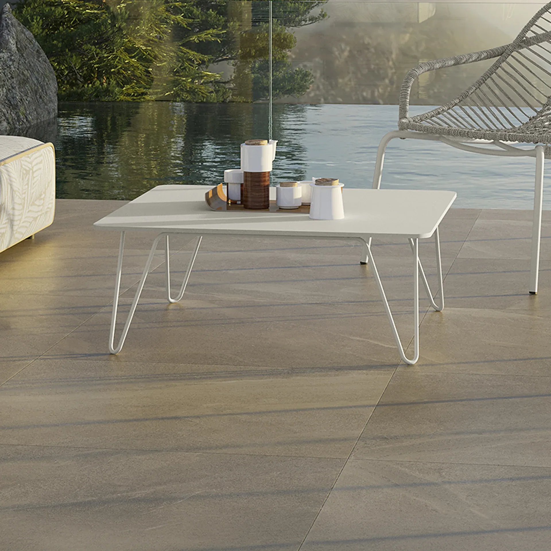 Outdoor patio with a white coffee table, coffee set, part of a sofa and chair, overlooking water with trees and a rock.