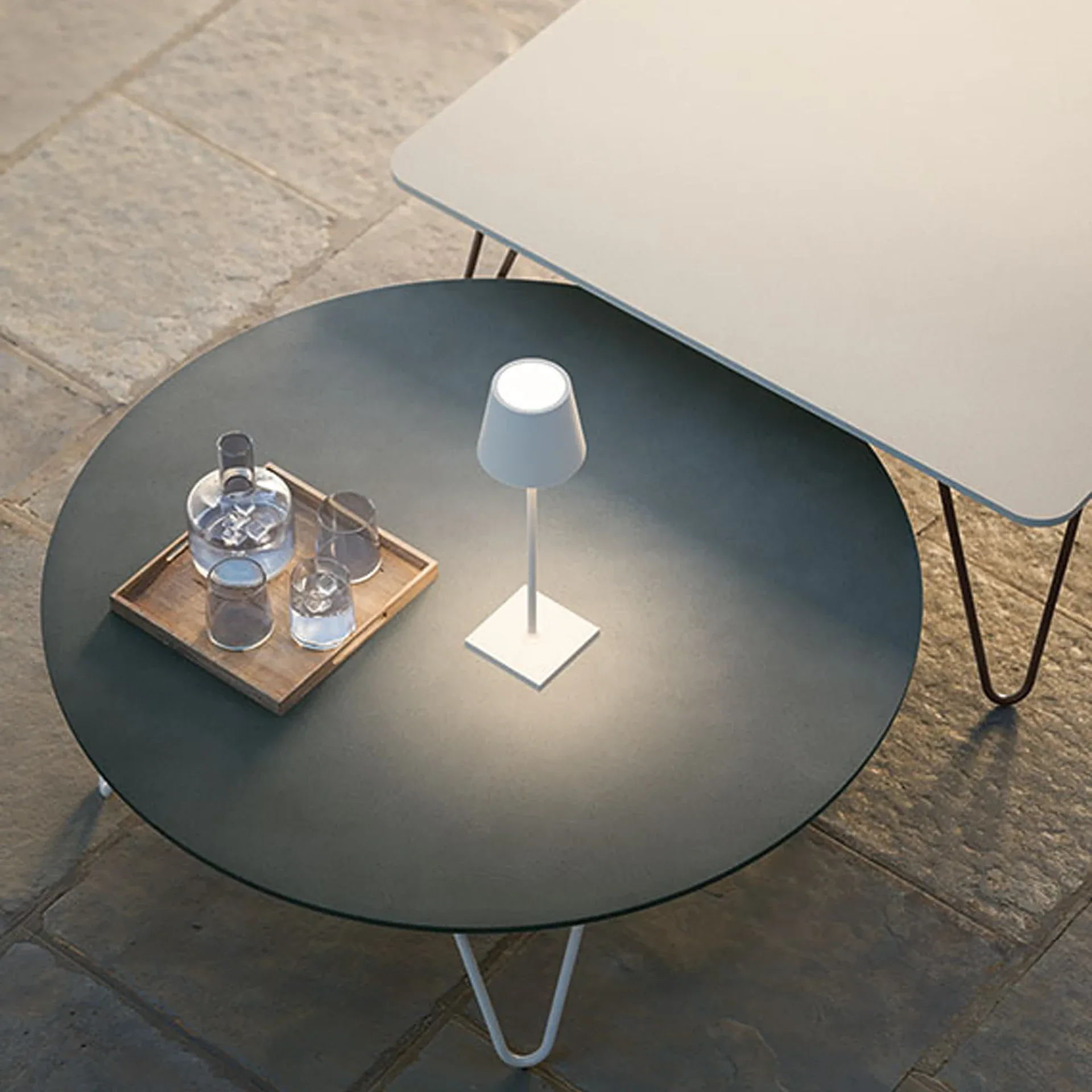 Overhead view of a dark grey round table and a light grey rectangular table on a stone patio. The round table has a lit white lamp and a tray with a glass decanter and glasses.