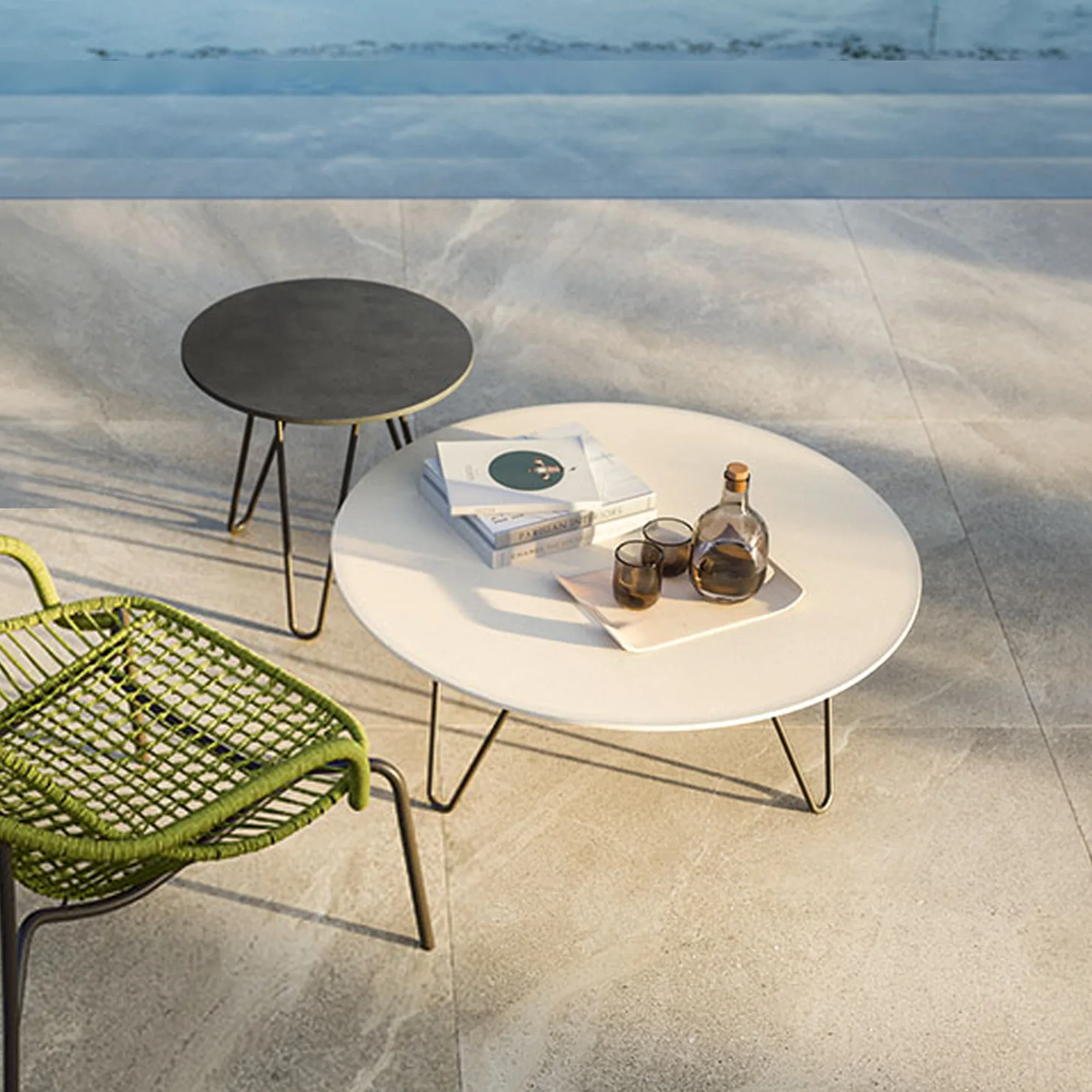 An outdoor patio with a round light beige coffee table, a smaller dark gray side table, and a lime green woven chair. The coffee table holds books, a decanter, and two glasses. Light-colored tiles cover the floor, with blurred blue water in the background.