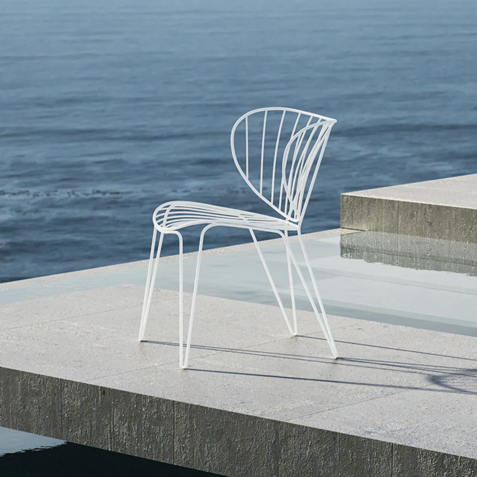 A white slatted wire chair on a light gray concrete patio overlooking calm blue water.