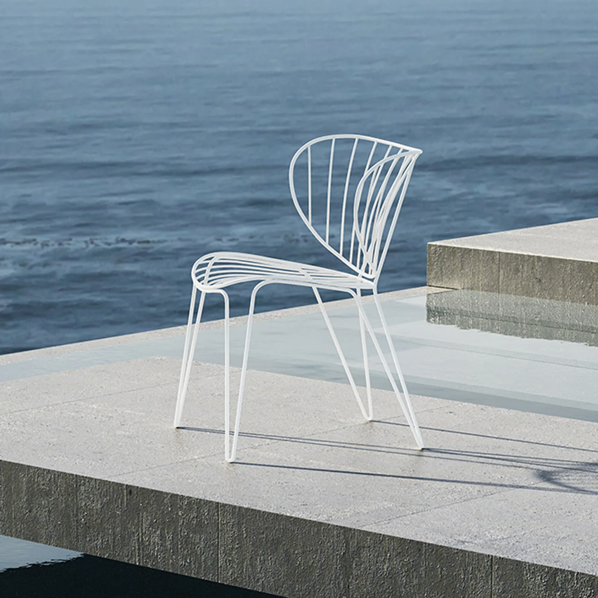A white slatted wire chair on a light gray concrete patio overlooking calm blue water.