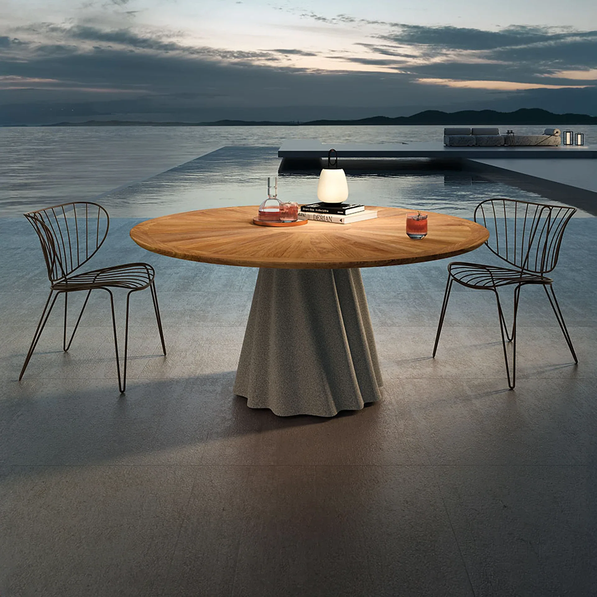 An outdoor dining table with two wireframe chairs by an infinity pool overlooking a body of water at dusk. A glowing lantern, books, and drinks are on the table.