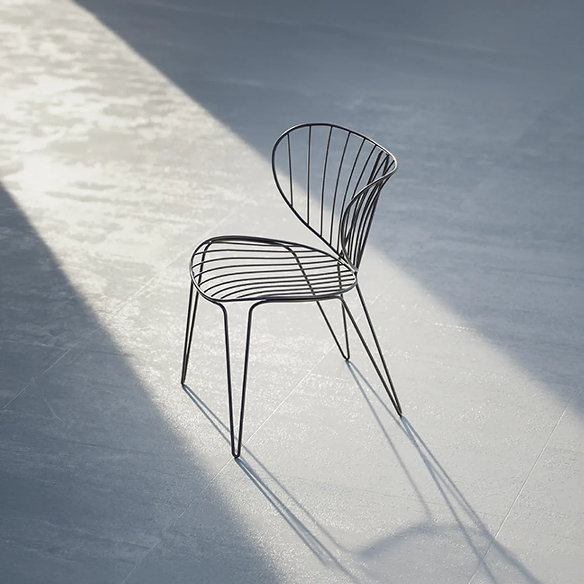 A black wire chair with a curved back and seat, standing on a light grey floor with sunlight and shadows.