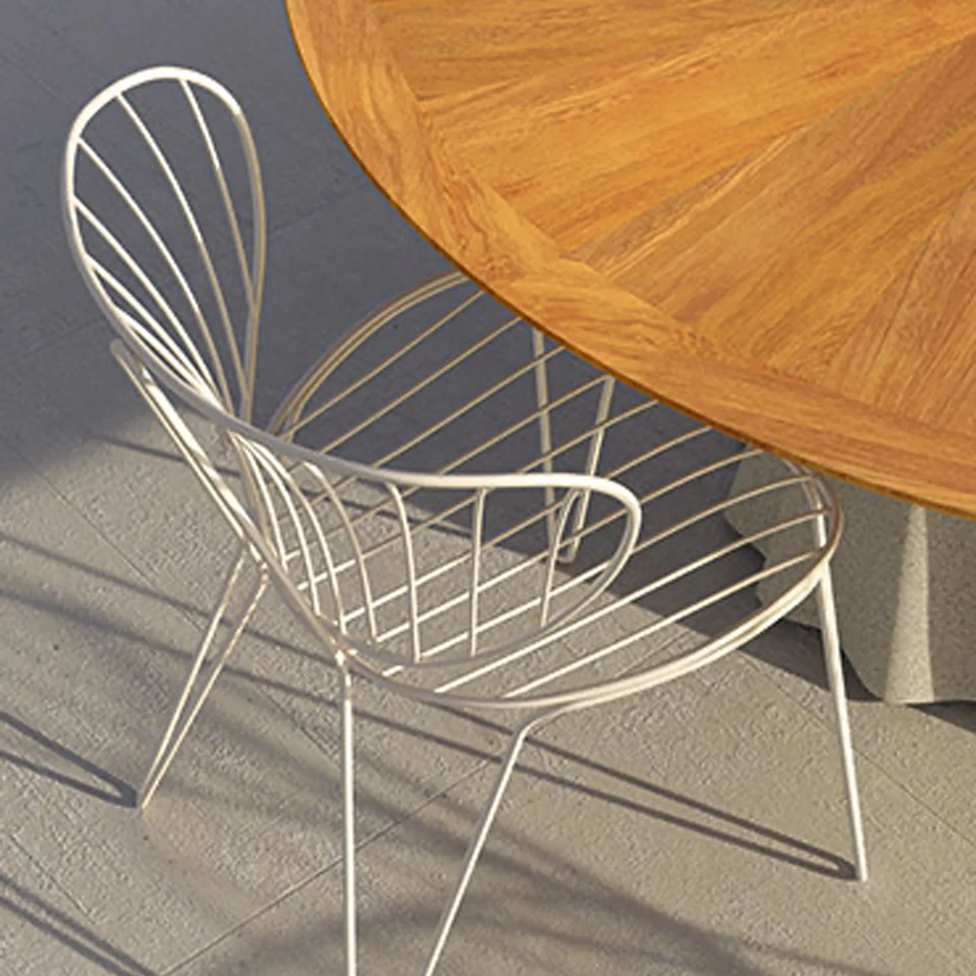 A white wire-frame chair partially under a round wooden table on a light gray textured floor, with strong shadows cast by the chair.