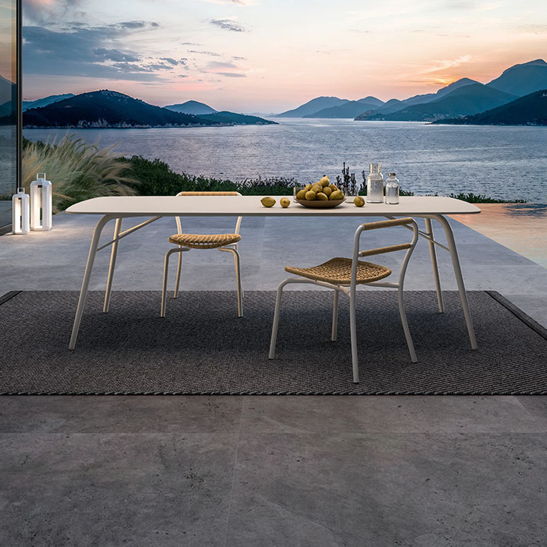 Outdoor patio with a light-colored dining table and two wicker chairs on a dark rug, overlooking a lake surrounded by mountains at sunset. The table holds fruit and glass bottles.