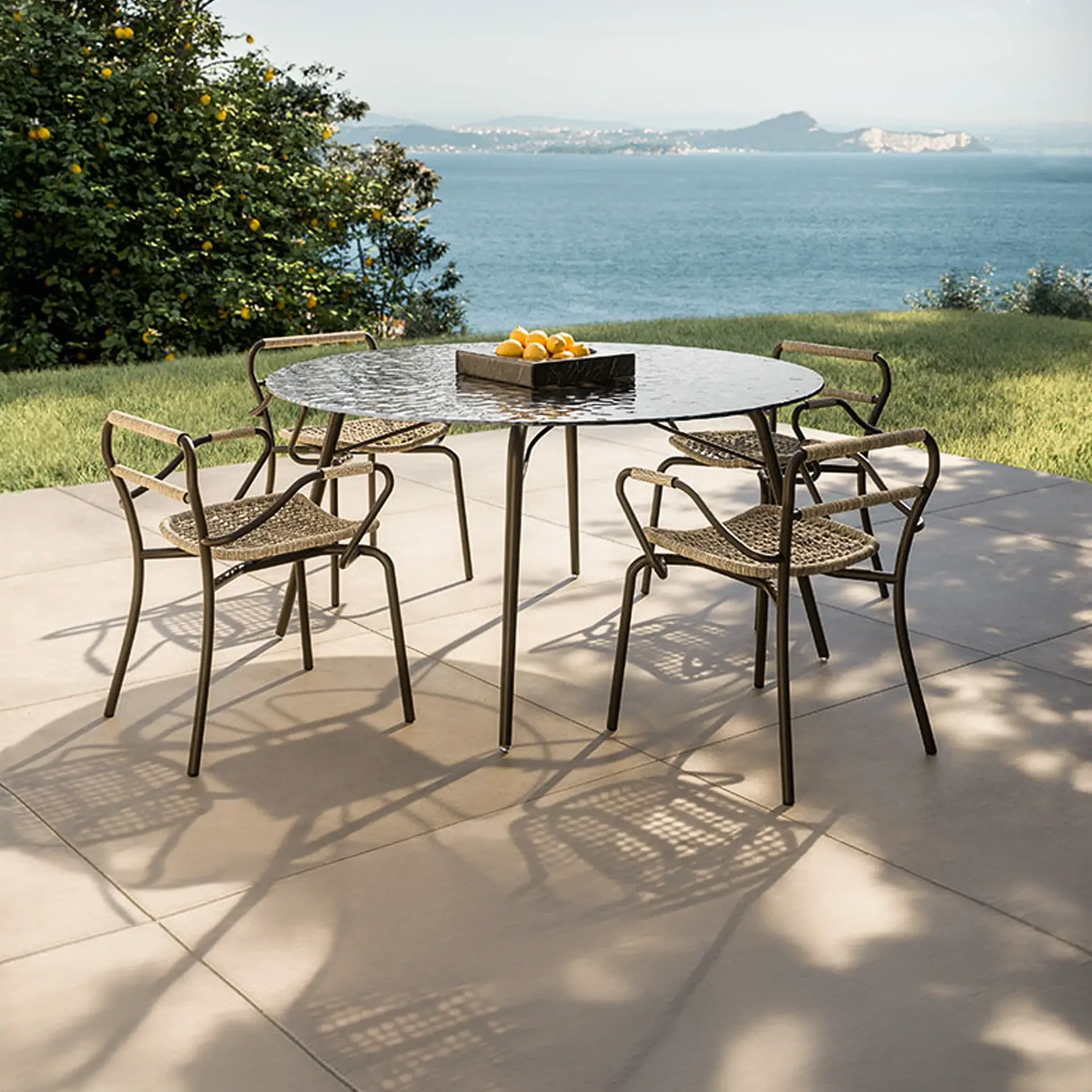 An outdoor patio dining set with a round table and four chairs, featuring a bowl of lemons, overlooking a large body of water with distant mountains and a lemon tree.