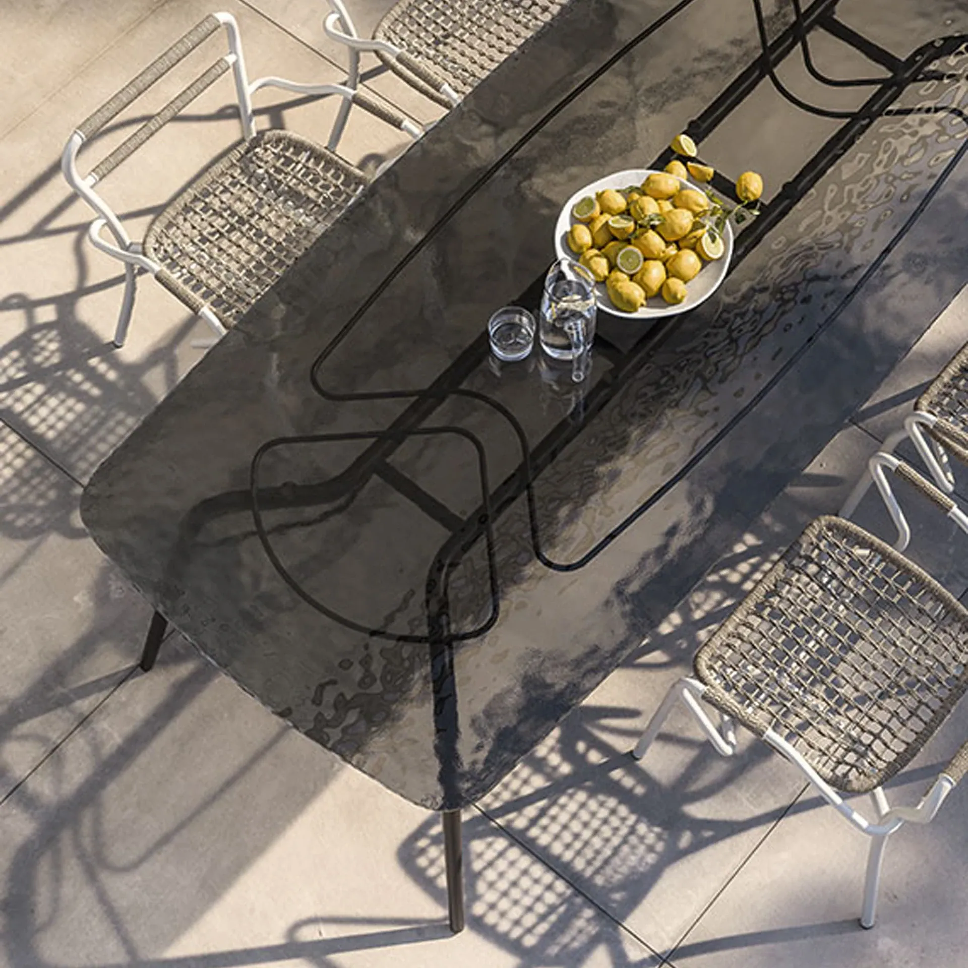 Overhead view of an outdoor dining table with a dark, reflective surface, surrounded by chairs with white frames and woven seats. A bowl of lemons, a water pitcher, and a glass are on the table. Strong shadows are cast on the patio.