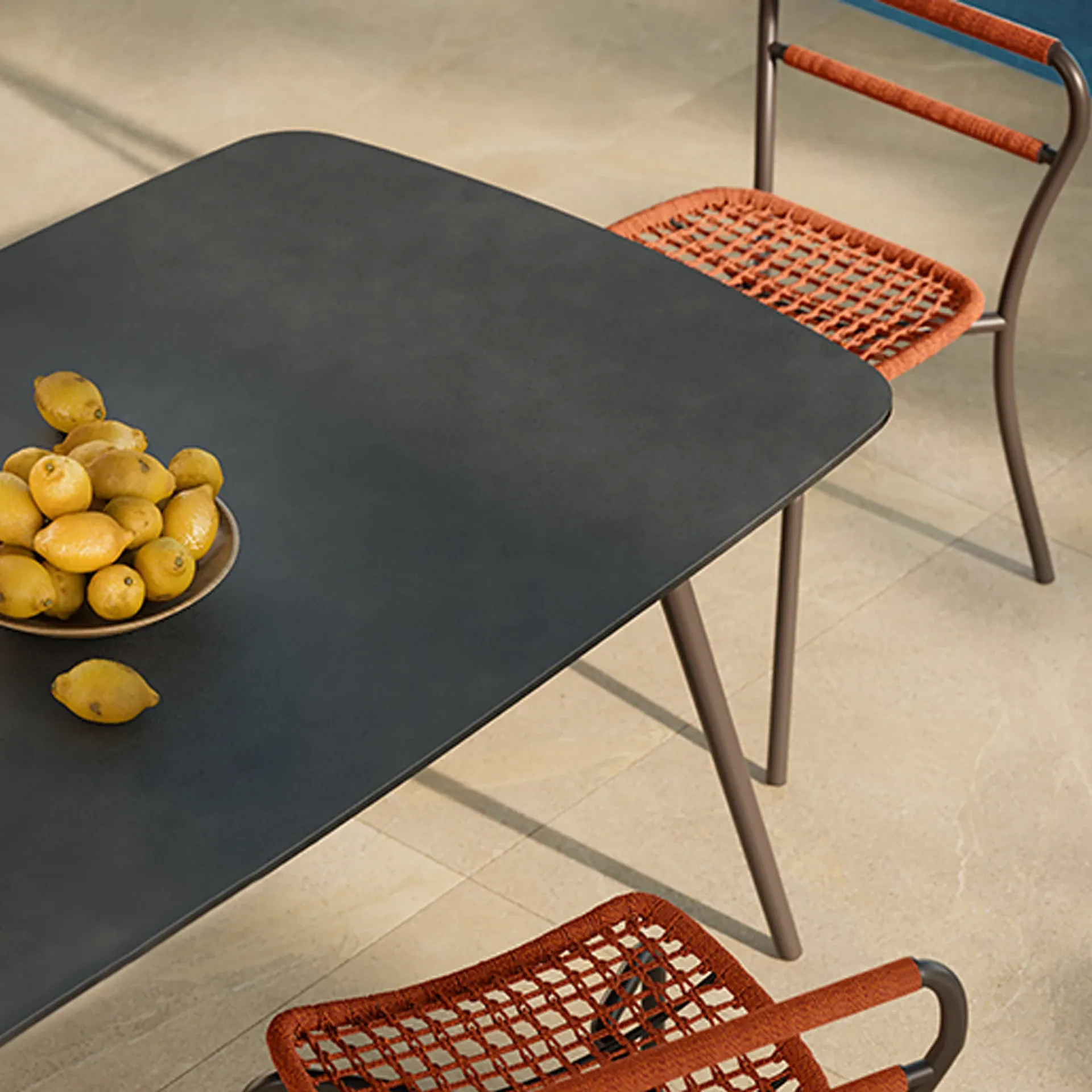 Overhead view of a dark gray table with a bowl of lemons, flanked by two chairs with orange woven seats, on a light tiled patio.