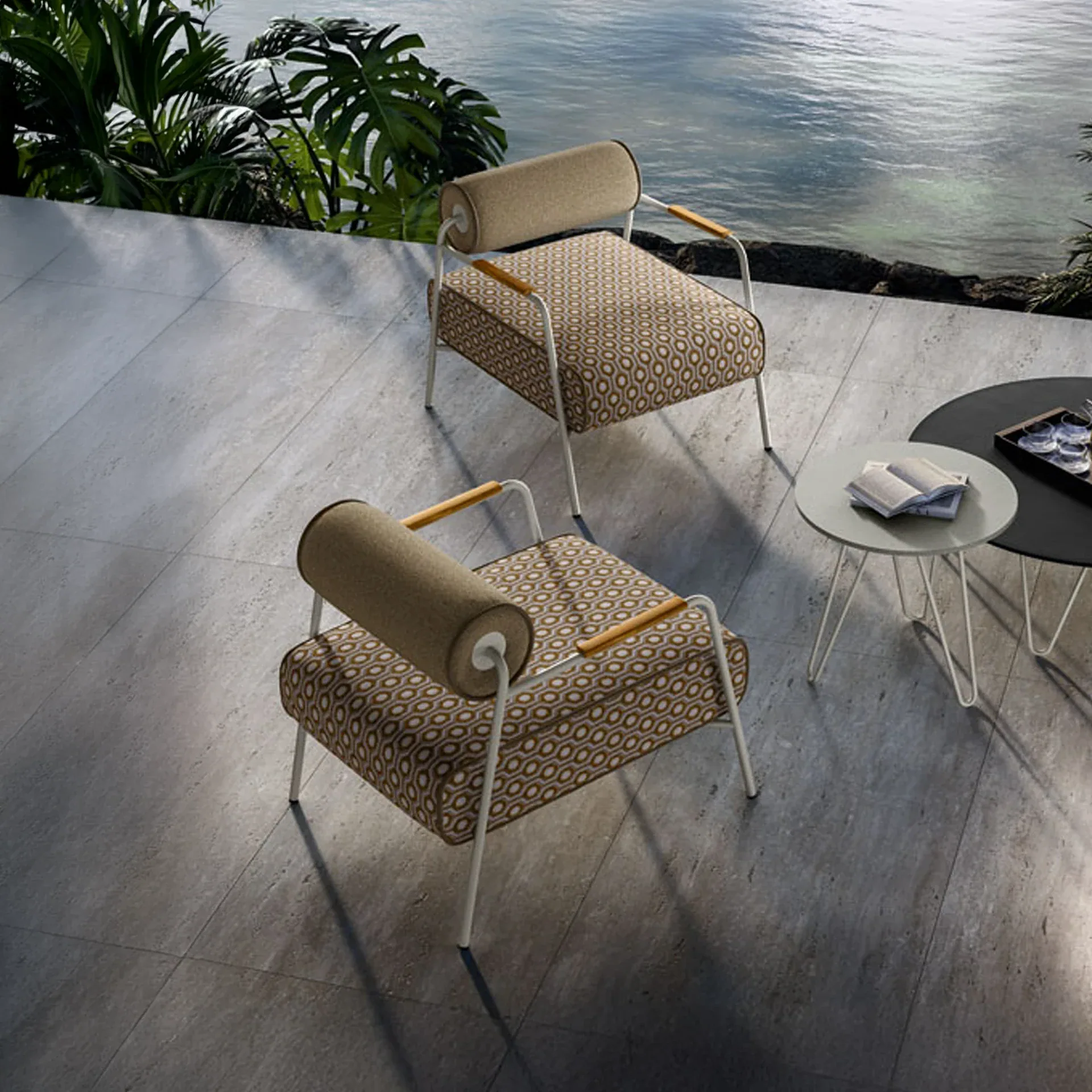 Two modern outdoor chairs with patterned cushions and two round tables on a grey tiled patio overlooking calm blue water with green foliage.