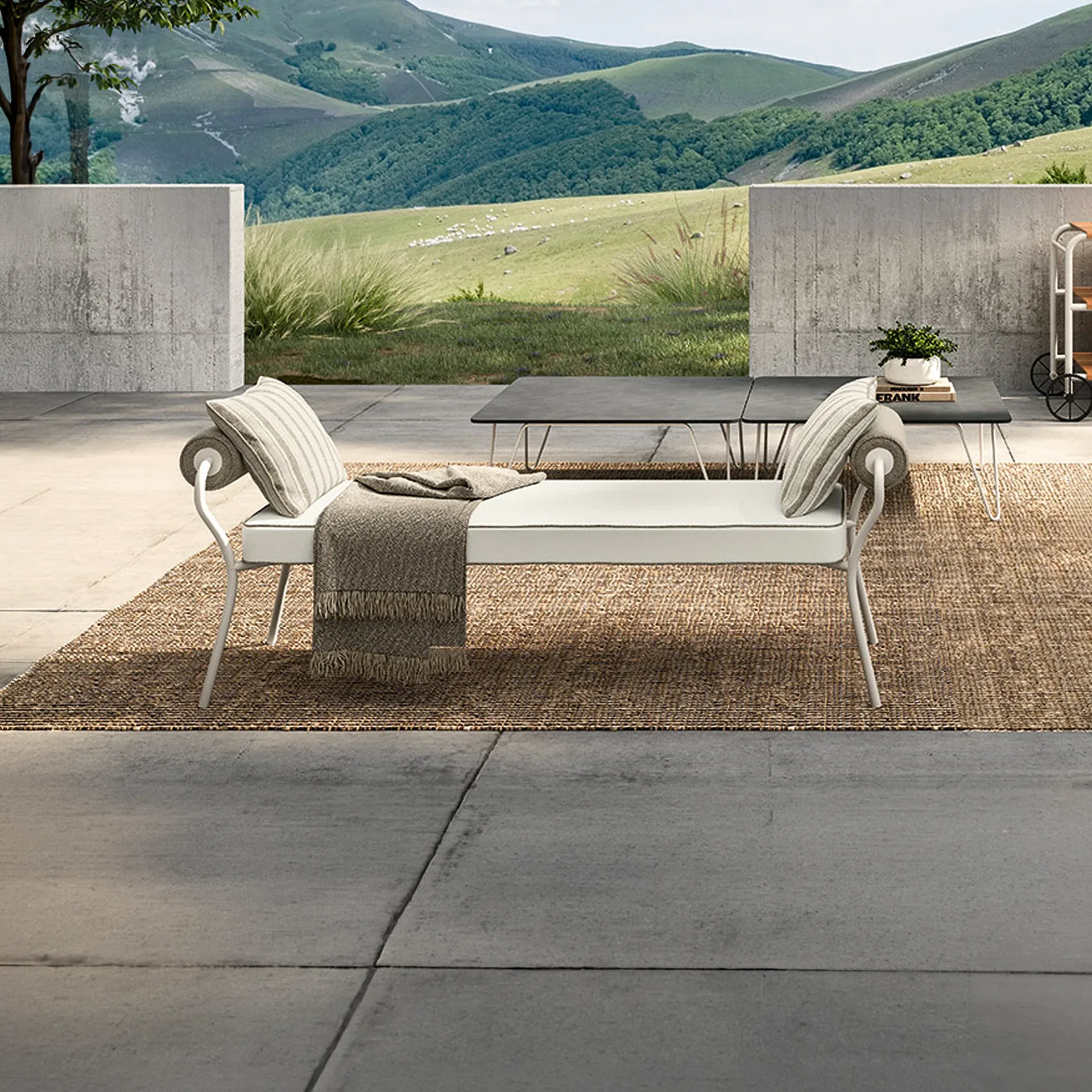 An outdoor patio with a white daybed, coffee tables, and a woven rug, set against a backdrop of rolling green hills.