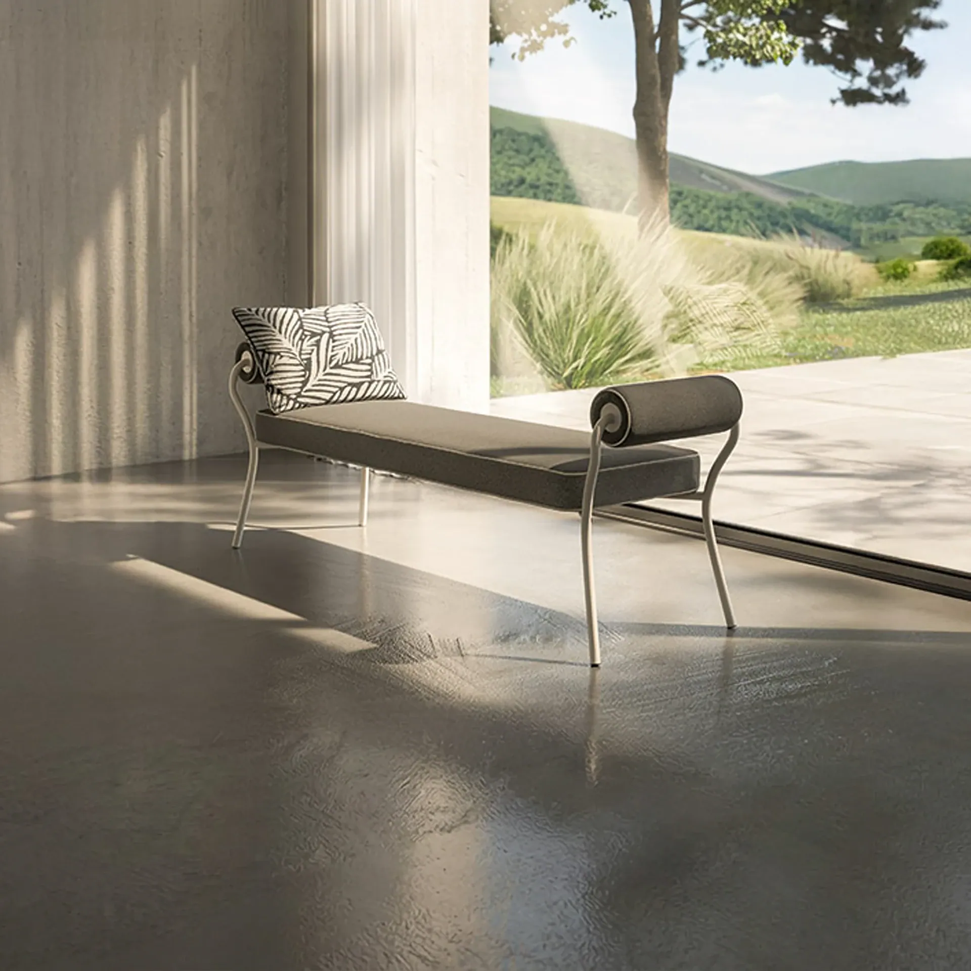 A modern dark gray bench with a patterned pillow sits on a polished concrete floor next to a large window overlooking a landscape of green hills, a tree, and ornamental grasses.