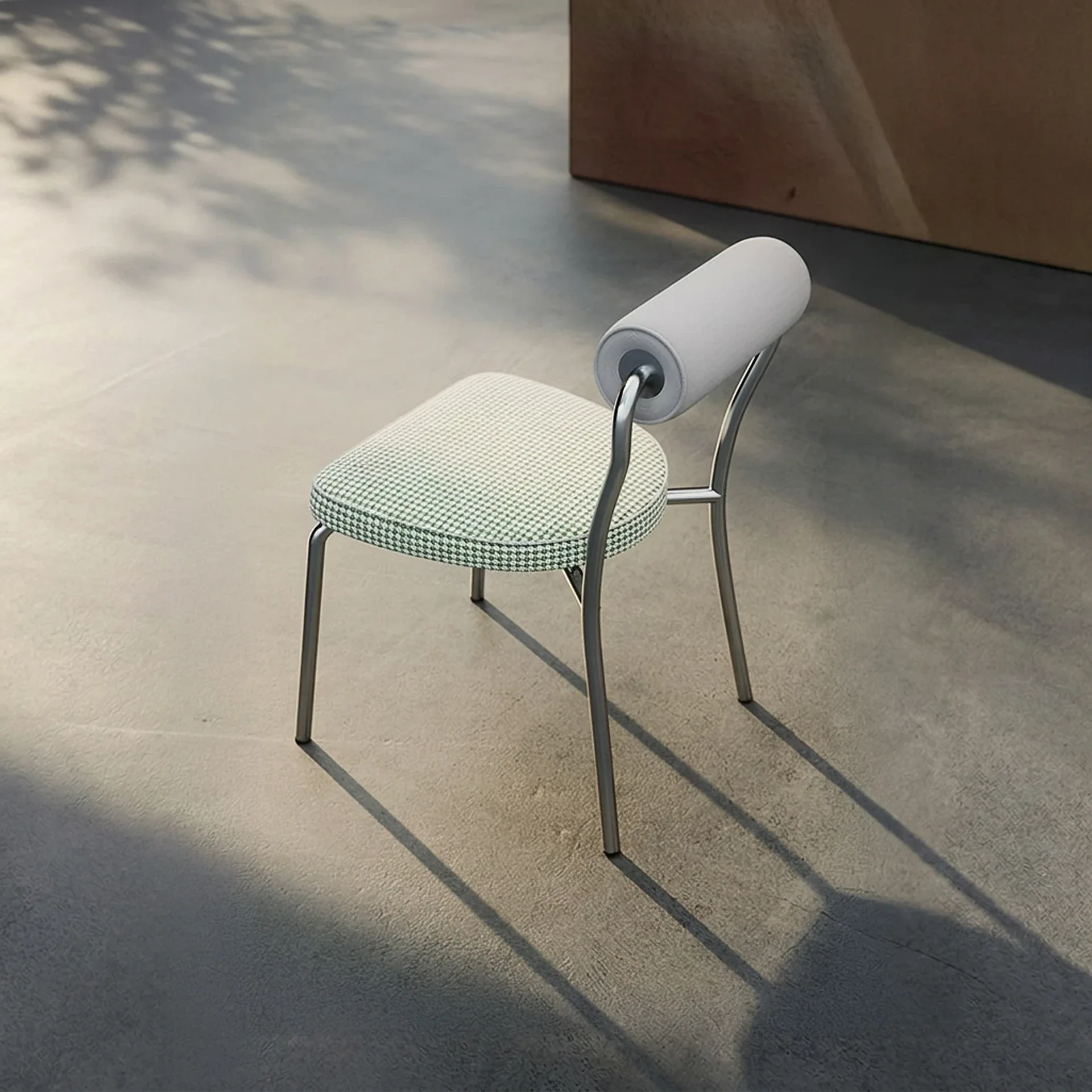 A modern chair with a silver frame, green and white patterned seat, and a cylindrical white backrest sits on a concrete floor with shadows.